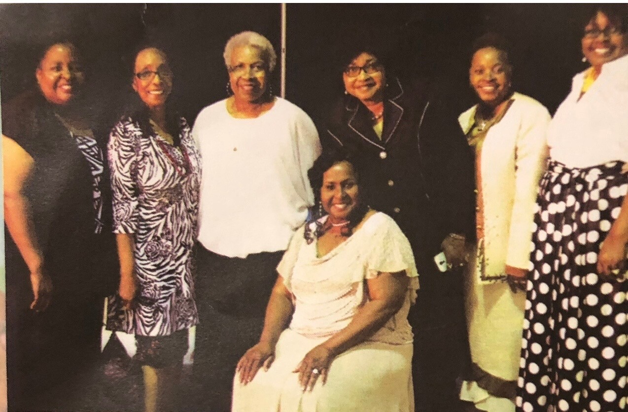 Seven women pose together for a group photo; the woman in white sits at center while others stand around her, smiling at an event.