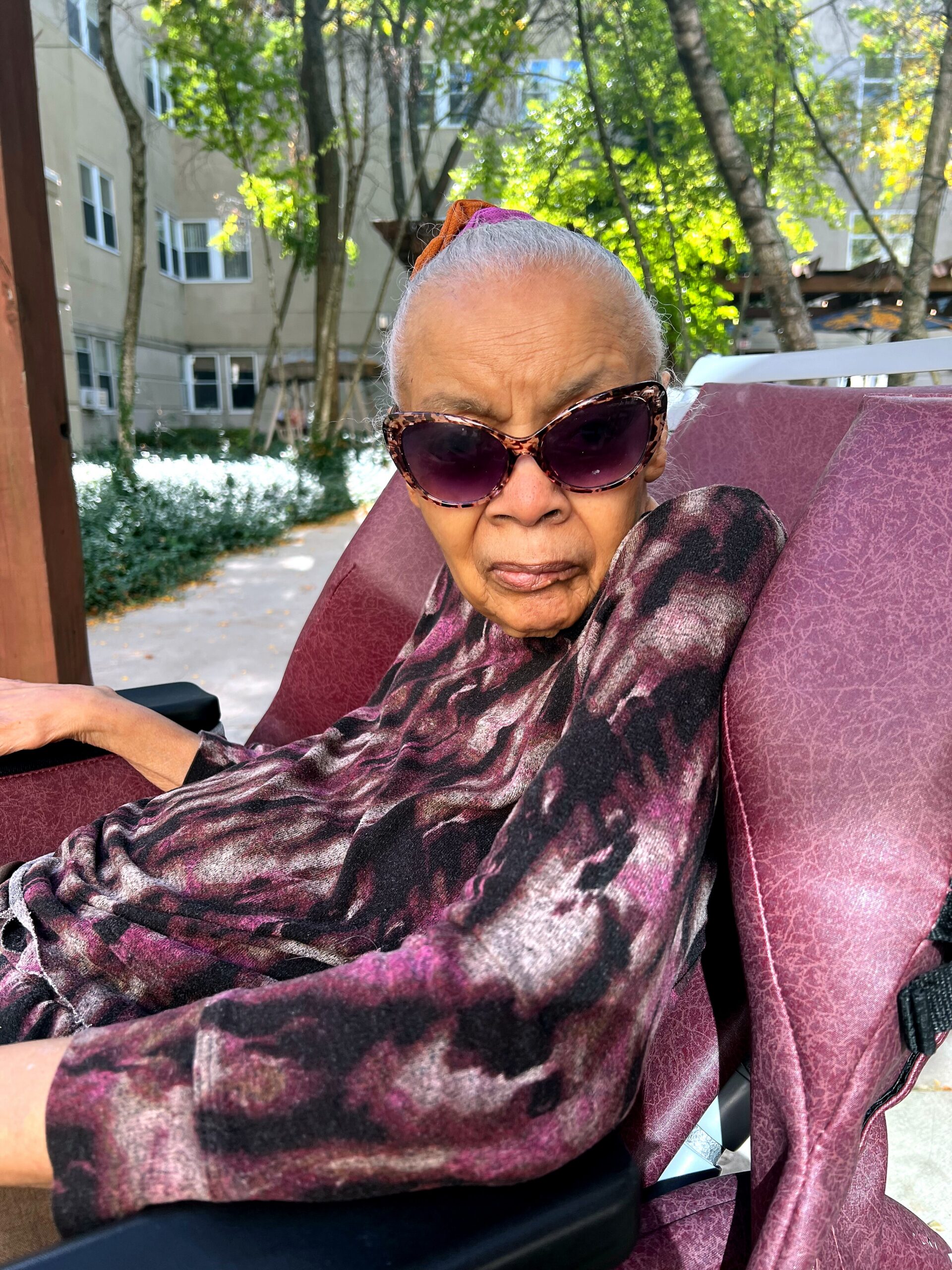 Older person wearing purple patterned shawl and tortoiseshell sunglasses sits in a maroon outdoor chair; trees and apartment buildings in the background.