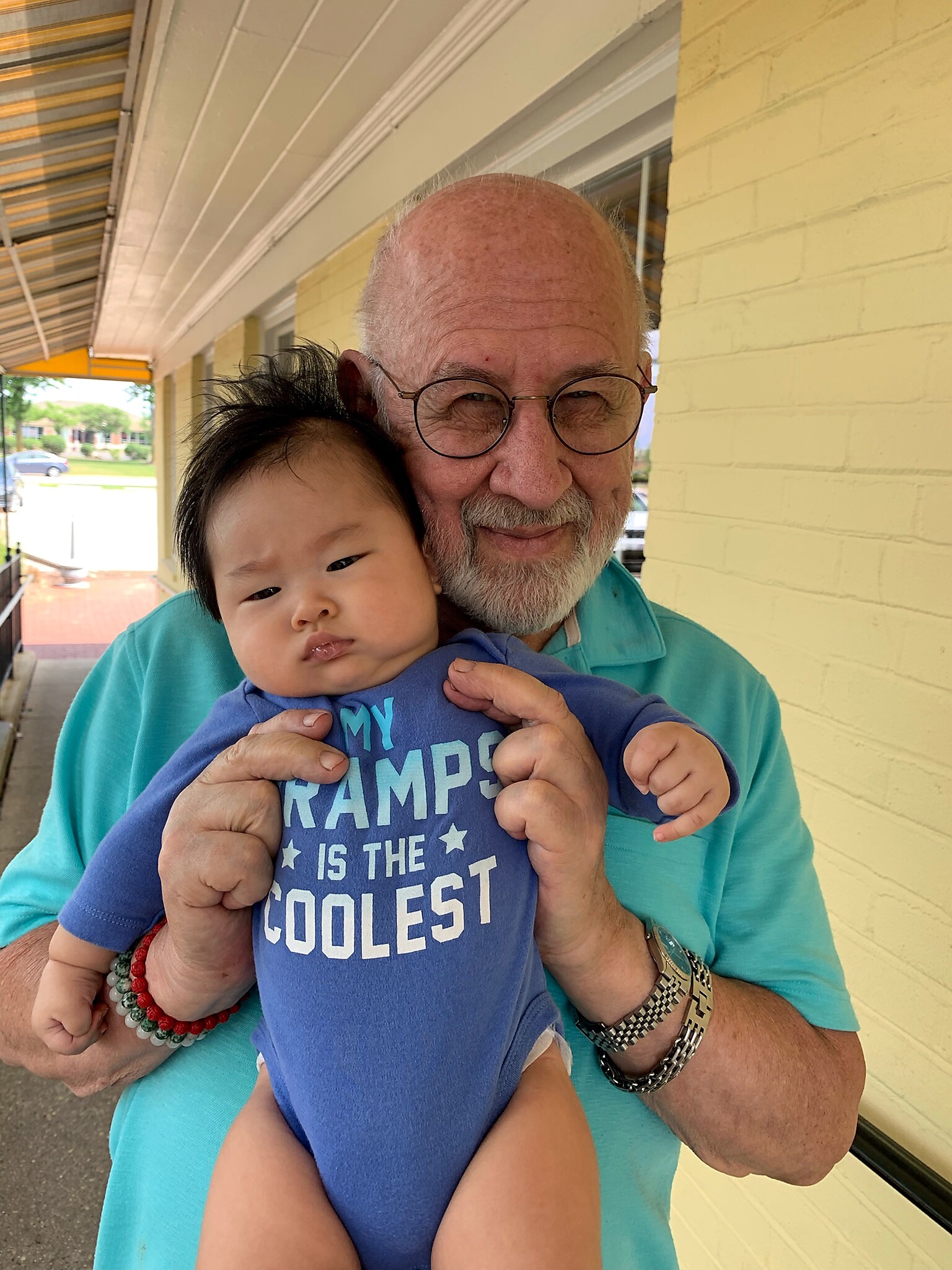 Senior man with glasses holding a baby close, both smiling at the camera, standing outdoors by a yellow brick wall.