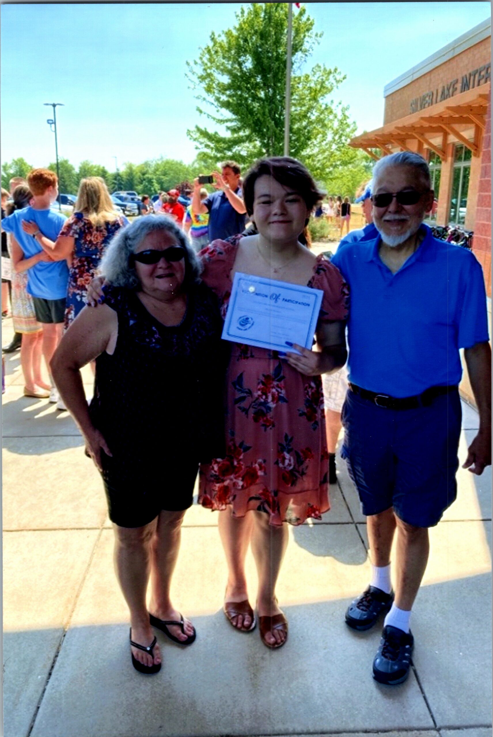 Three adults pose outdoors; center woman in a floral dress holds a certificate, flanked by a woman in black and a man in blue.