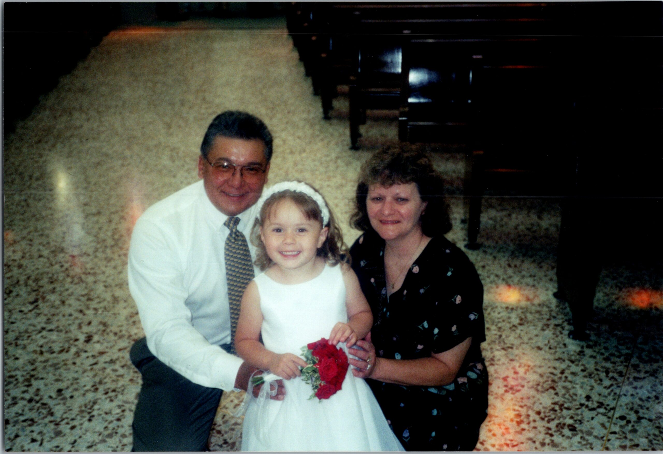 Smiling family posing outdoors: father in a white shirt and tie, daughter in a white dress holding a red bouquet, mother in a dark dress beside them.