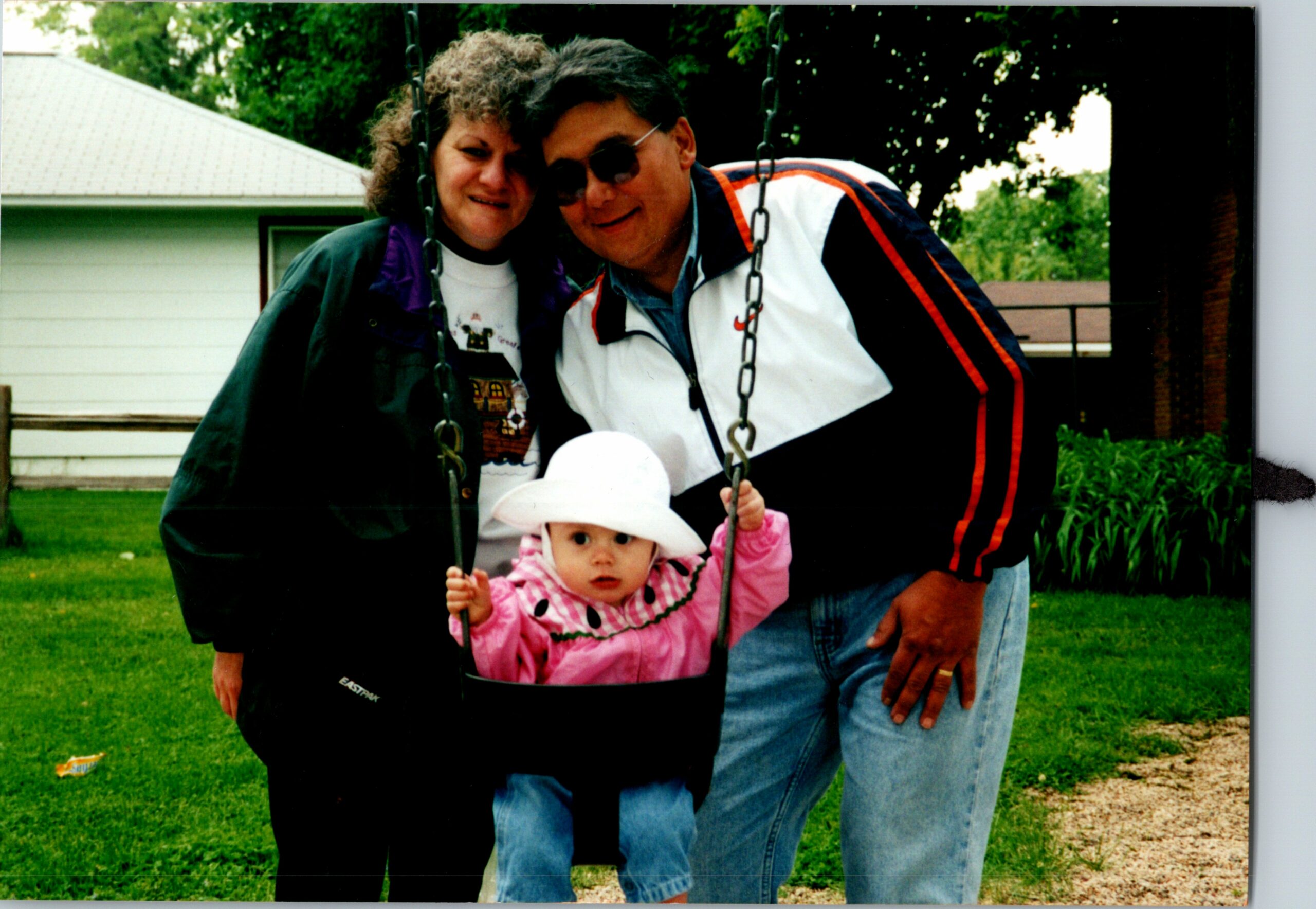 Older couple smiling beside their baby daughter in a pink jacket on a swing in a backyard. Green lawn, trees, and a house in the background.