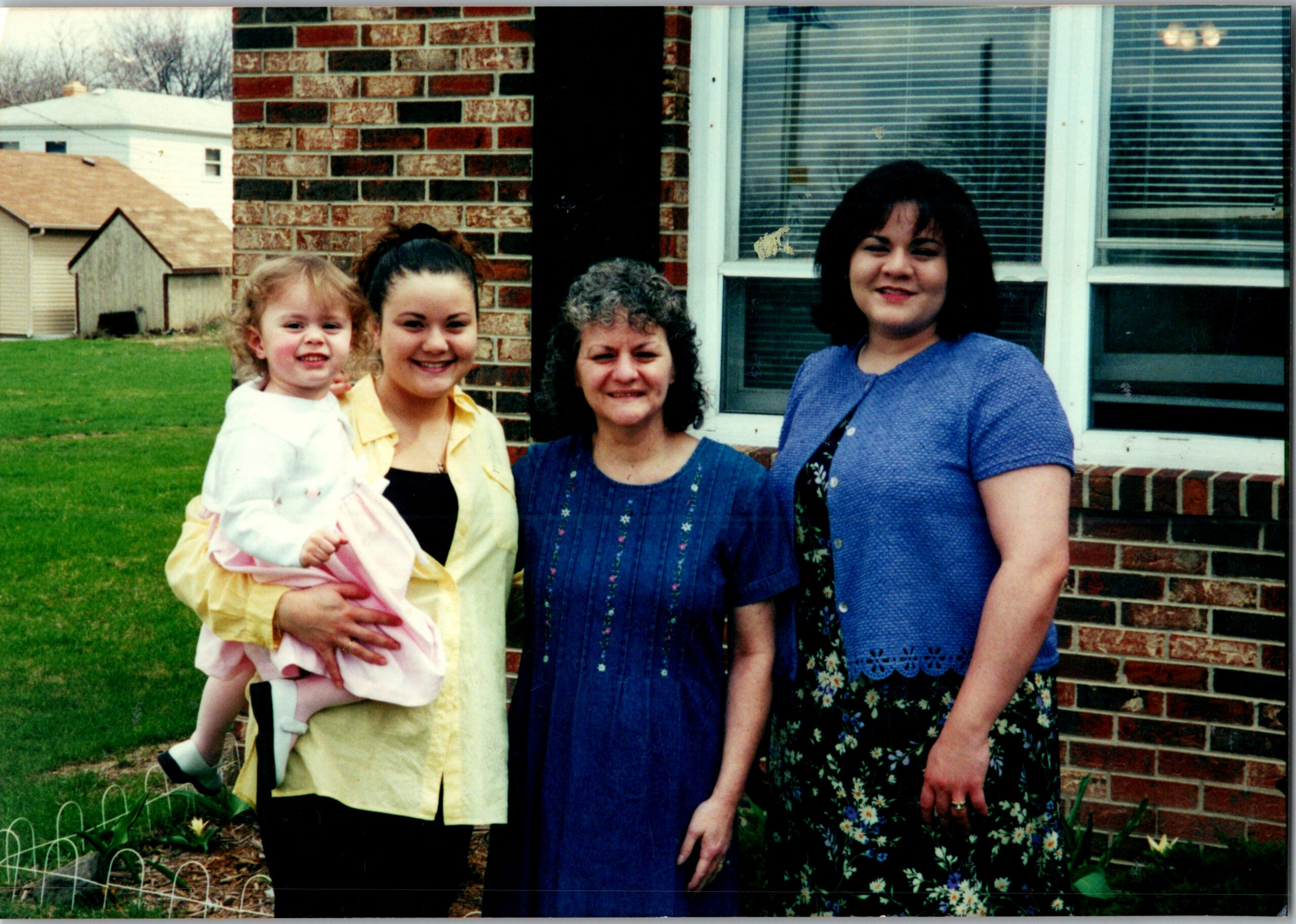 Four women pose in a backyard beside a brick house; a young girl is held by a woman in yellow, while the others wear blue dresses, all smiling.