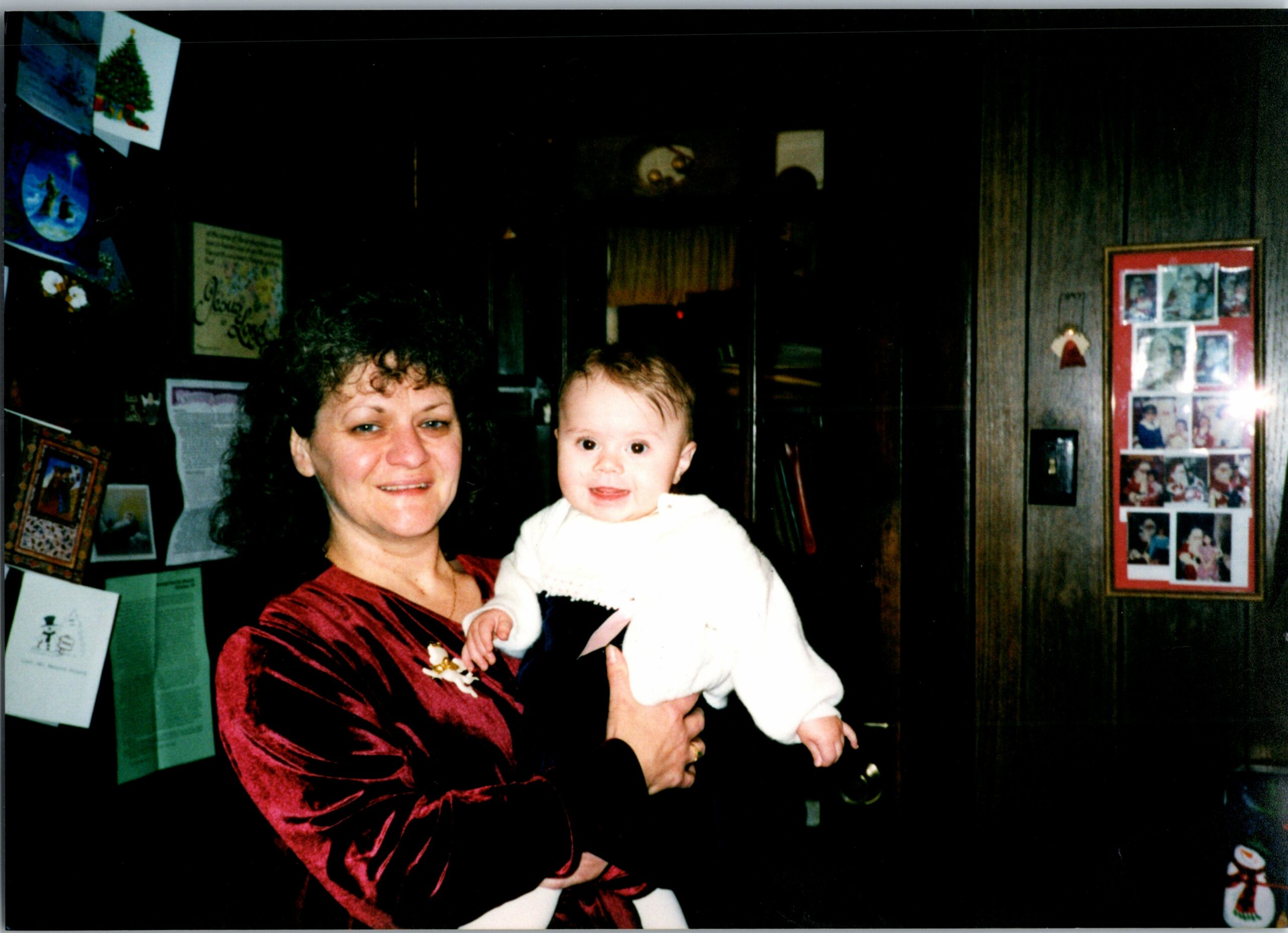 Woman in a red top holds a smiling baby indoors, with holiday cards and photos on the wall behind them.
