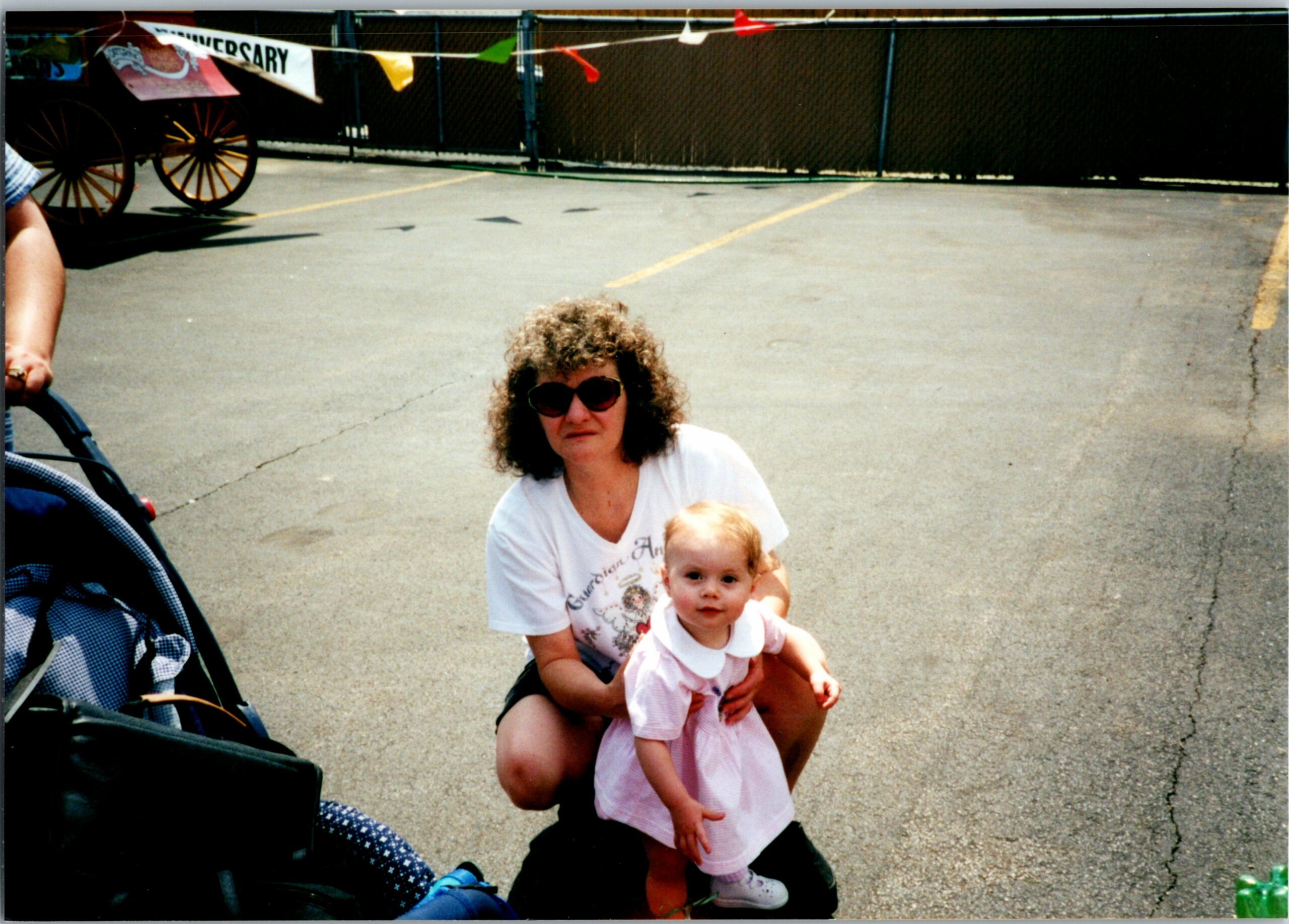 Woman wearing sunglasses kneels in a parking lot, holding a baby in a pink dress; a stroller is visible on the left.