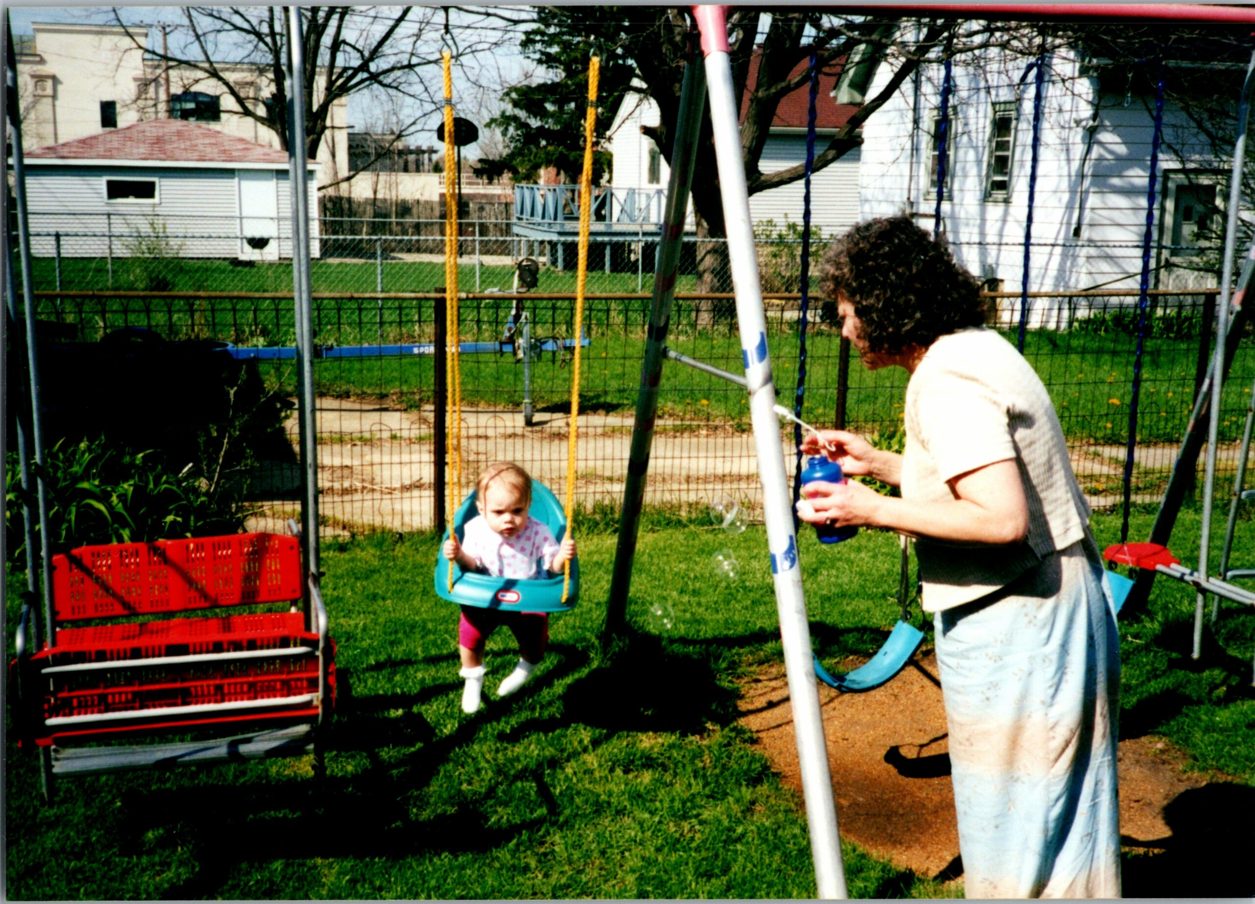 A woman holding a water bottle standing beside a child in a swing set in a sunny backyard.