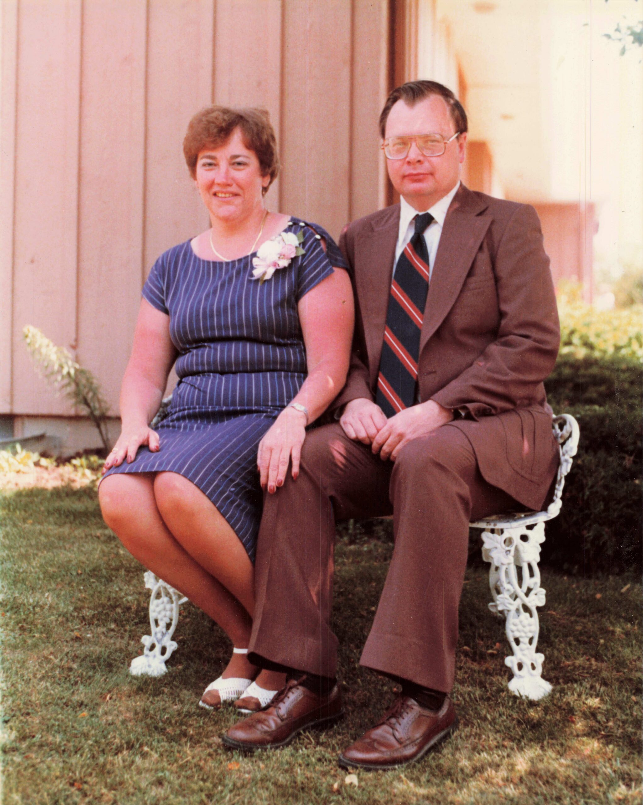 Couple sitting on a decorative white bench outdoors; woman in a blue striped dress with corsage, man in brown suit and striped tie.