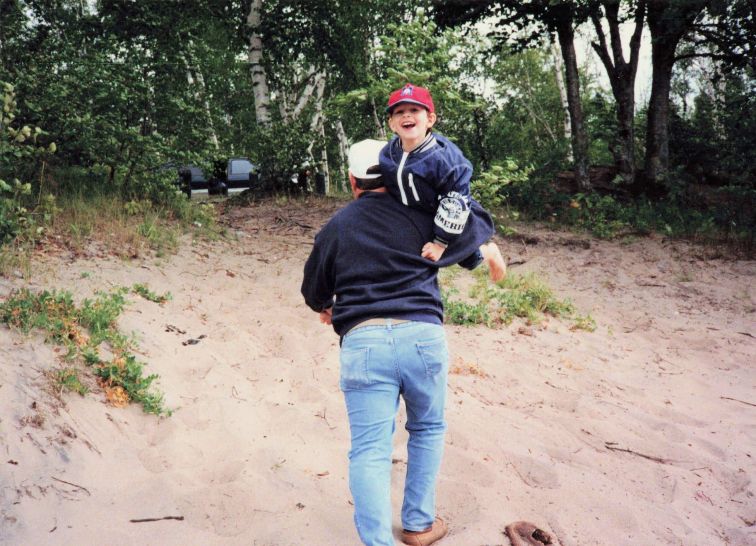 Young boy in a red cap riding on an adult's shoulders along a sandy, forested path, smiling.