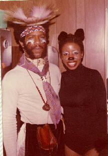 Two people in vintage costumes posing for a photo: man wearing a feathered headdress, beads, and sash beside a woman in a black bodysuit with buns.