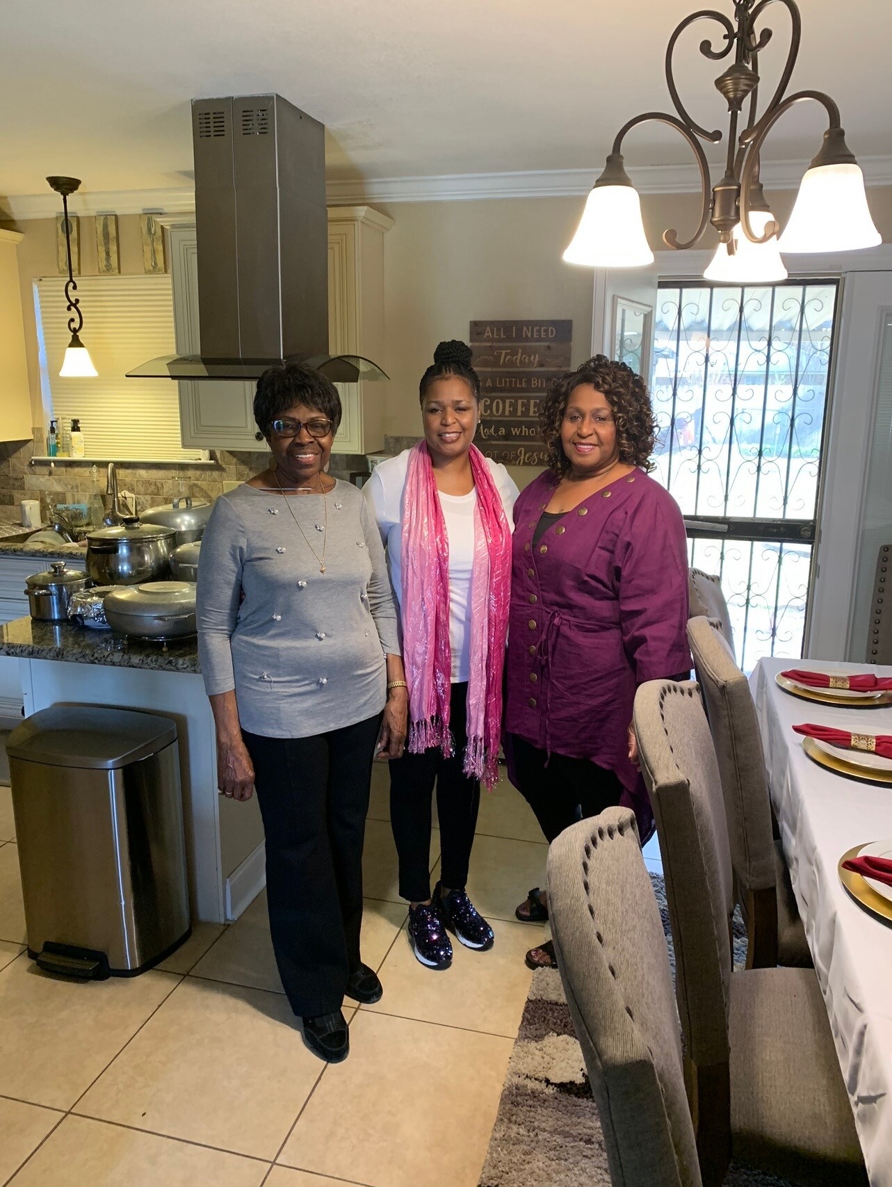 Three women stand together in a bright kitchen/dining area, smiling for the camera near a stove and dining table set for a meal.