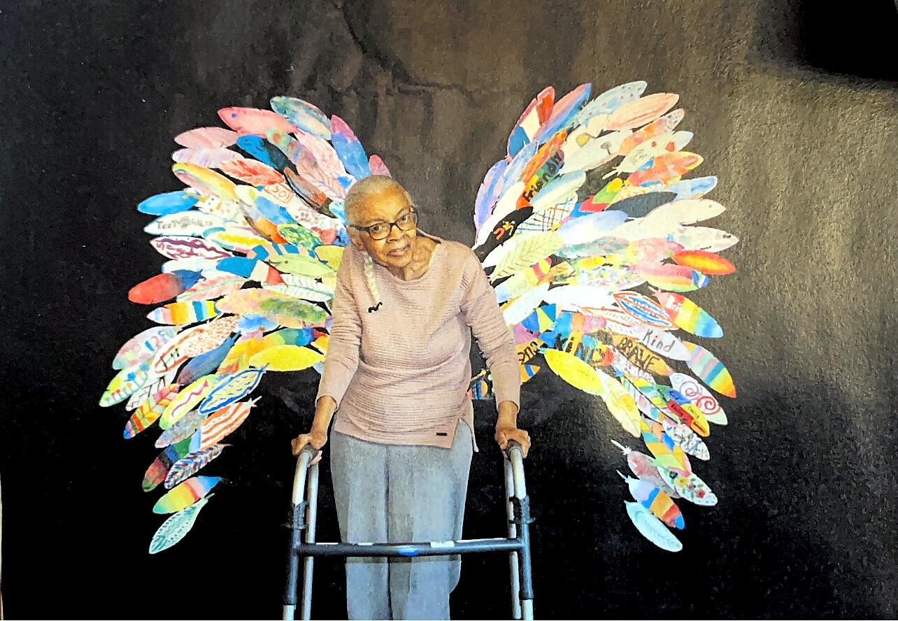 Elderly woman with glasses using a walker, standing before a wall mural of rainbow-colored paper wings.] ,