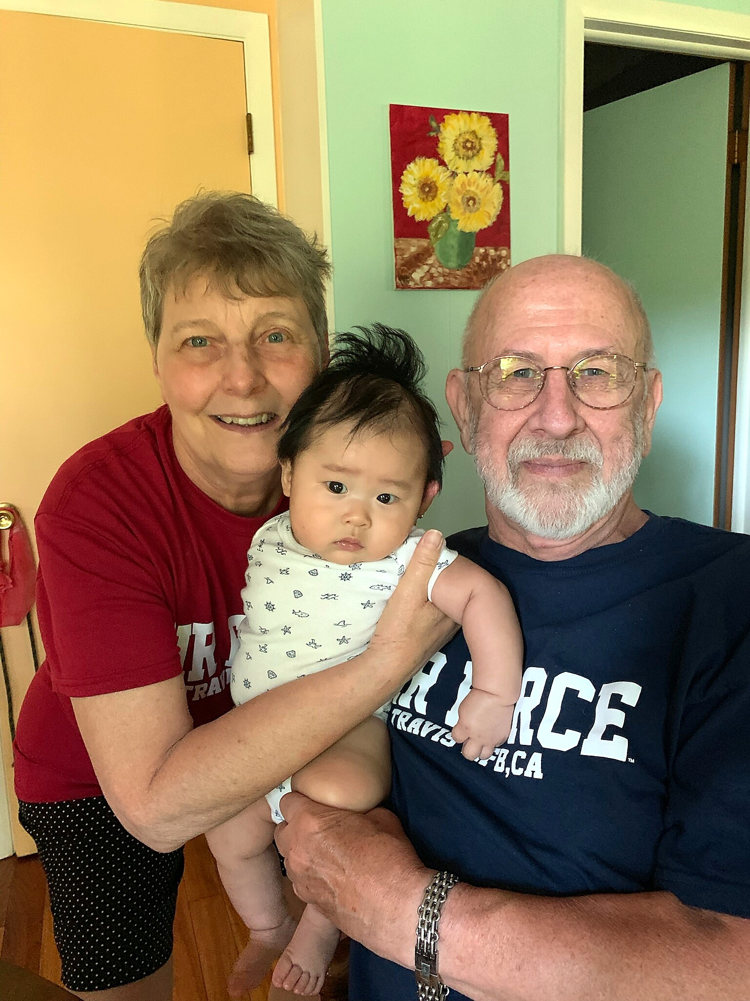 A smiling woman, an older man with glasses, and a baby pose for a selfie indoors, wearing casual clothes.