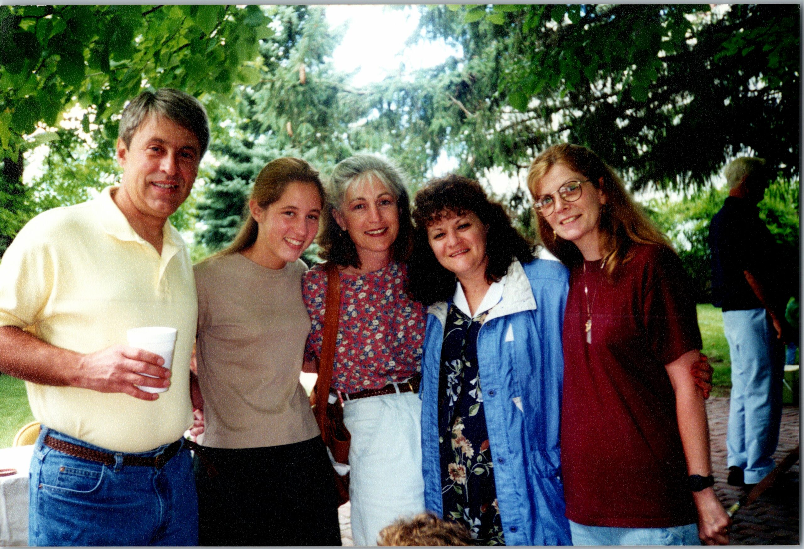 Six adults posing together outdoors in a park, smiling; man on the left holds a white cup.