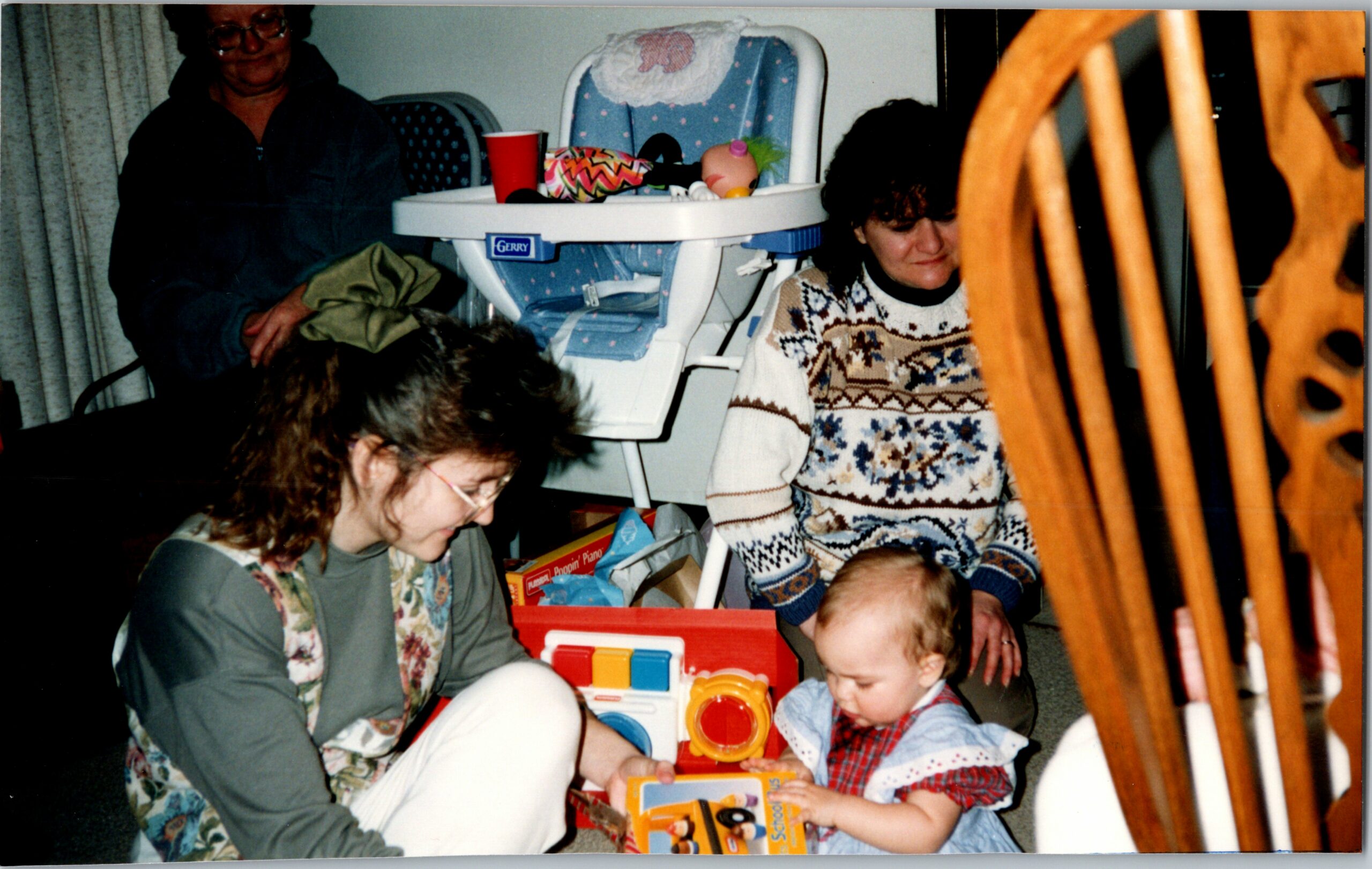 Family group in a living room with a baby and two women, a toddler looking at toys.