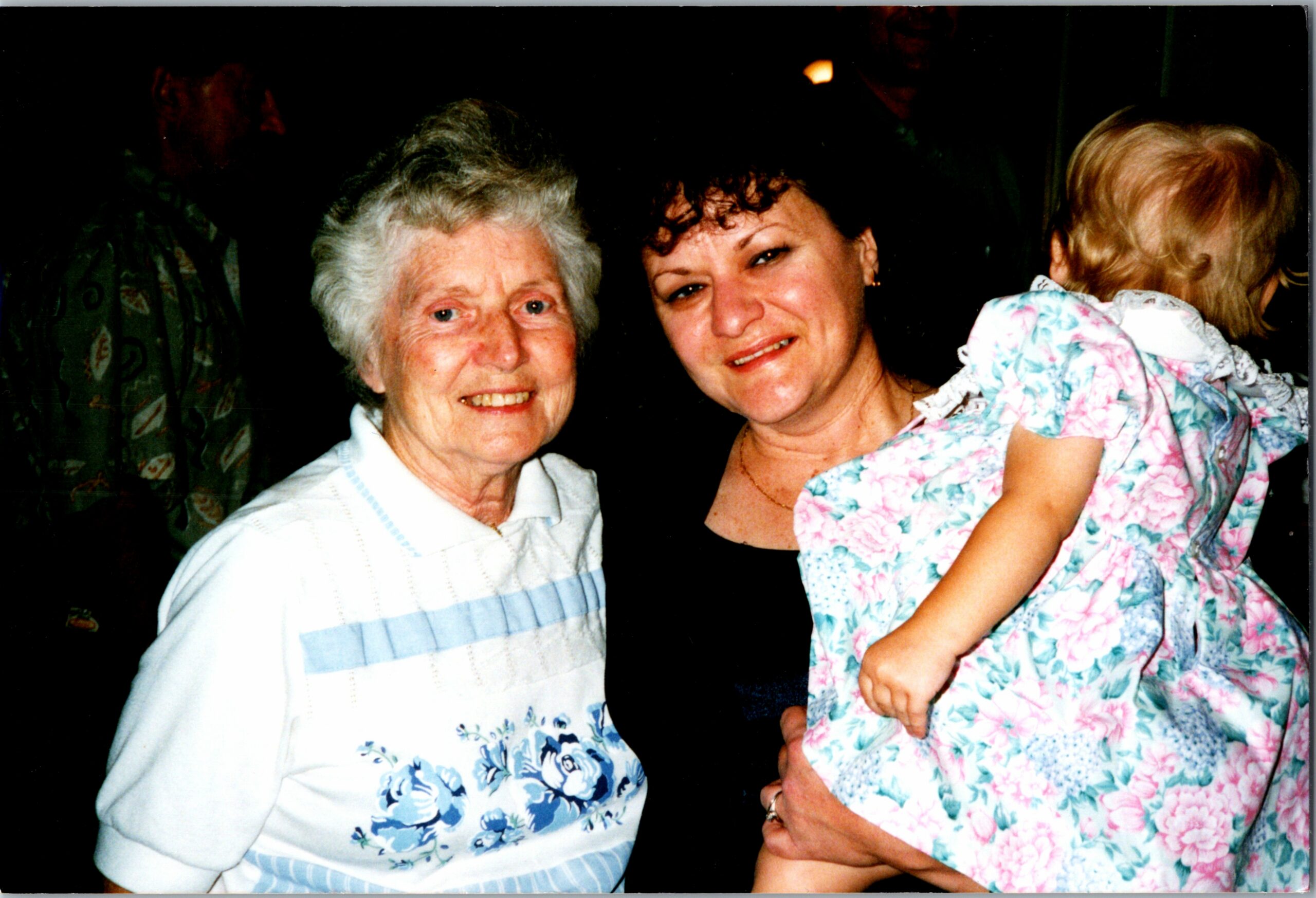 Three generations posing together: an elderly woman on the left, a middle-aged woman in the center, and a baby in a floral outfit on the right.