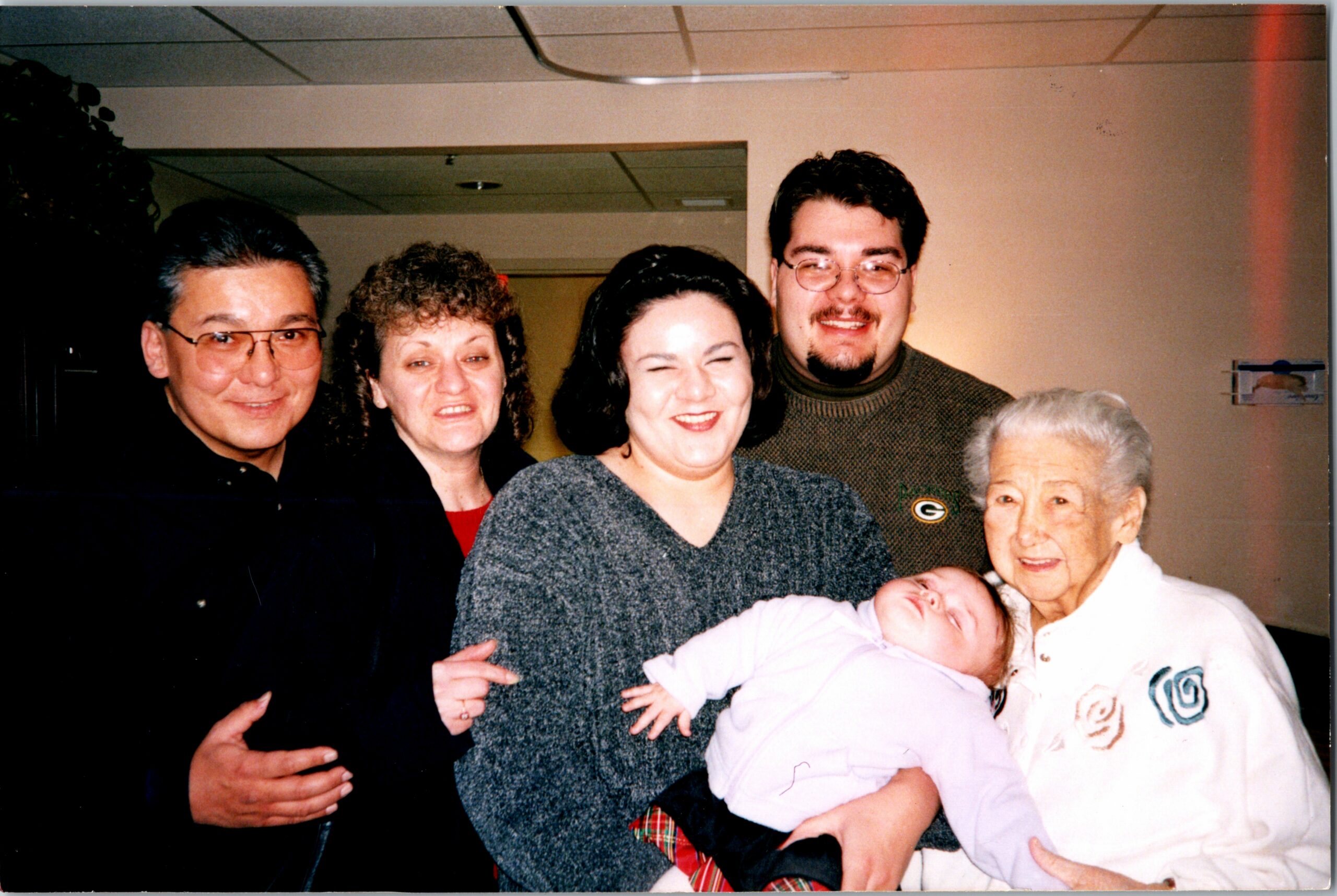 Multigenerational family portrait indoors: adults smile around a newborn baby being held by a woman in a gray sweater.