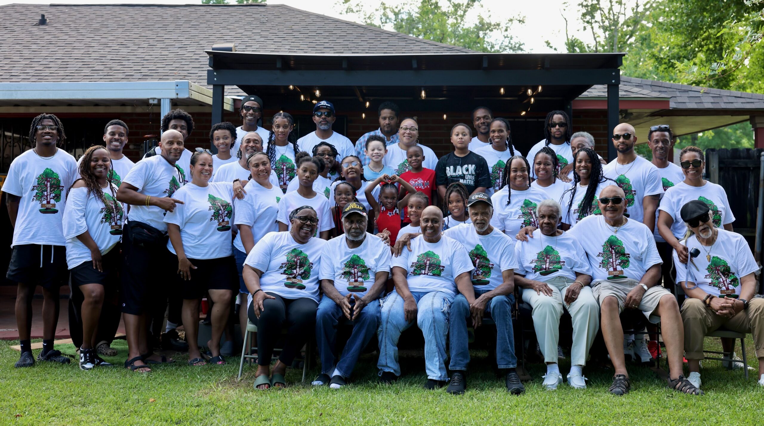 Large group of diverse people wearing matching white T-shirts with a tree graphic, posing on a backyard lawn.
