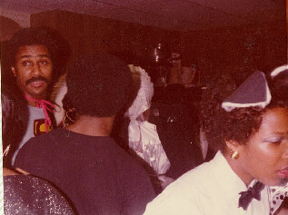 Group of people at a dimly lit party; a man with an afro and mustache looks toward the camera on the left while others mingle in the background.