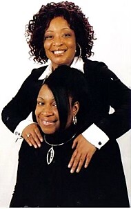 Two women smiling and posing in a studio portrait, with one standing behind the other and resting hands on her shoulders.