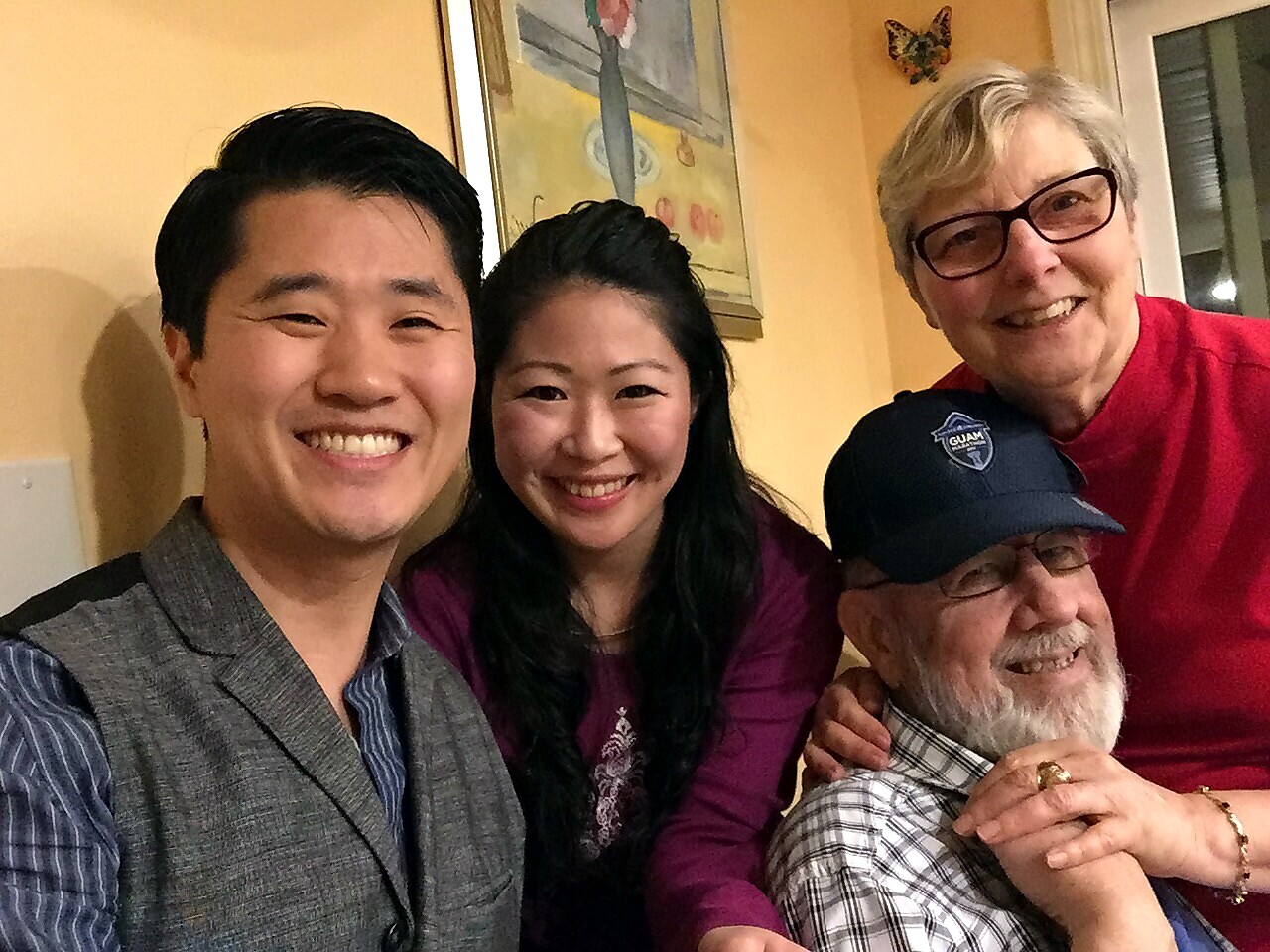 Four people indoors posing for a group selfie, smiling at the camera—two young adults on left, an older couple on the right.