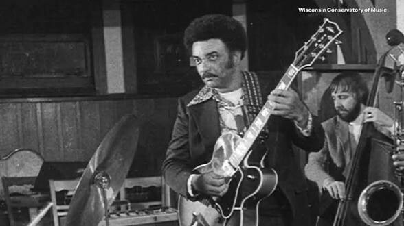 Black-and-white photo of a guitarist performing on stage with a bassist in the background; watermark reads Wisconsin Conservatory of Music.