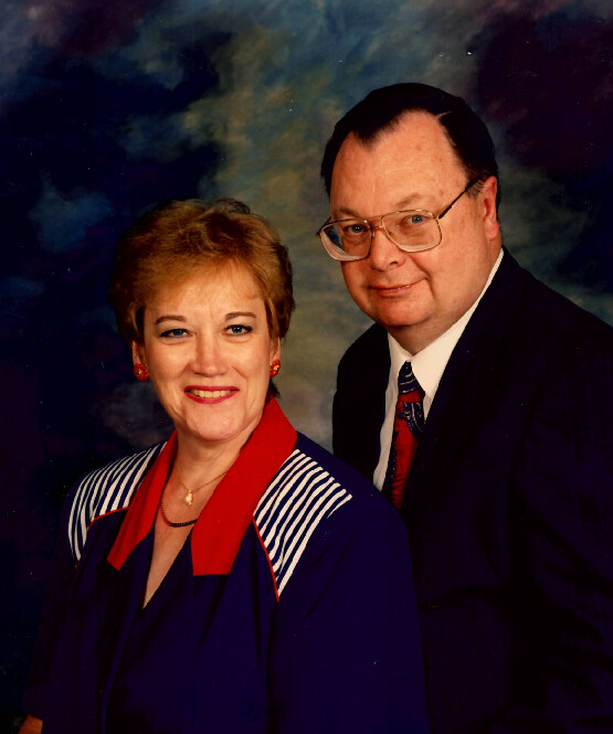 A smiling couple posing for a studio portrait: woman in a blue outfit with a red scarf and gold earrings, and a man in a dark suit and glasses standing beside her.