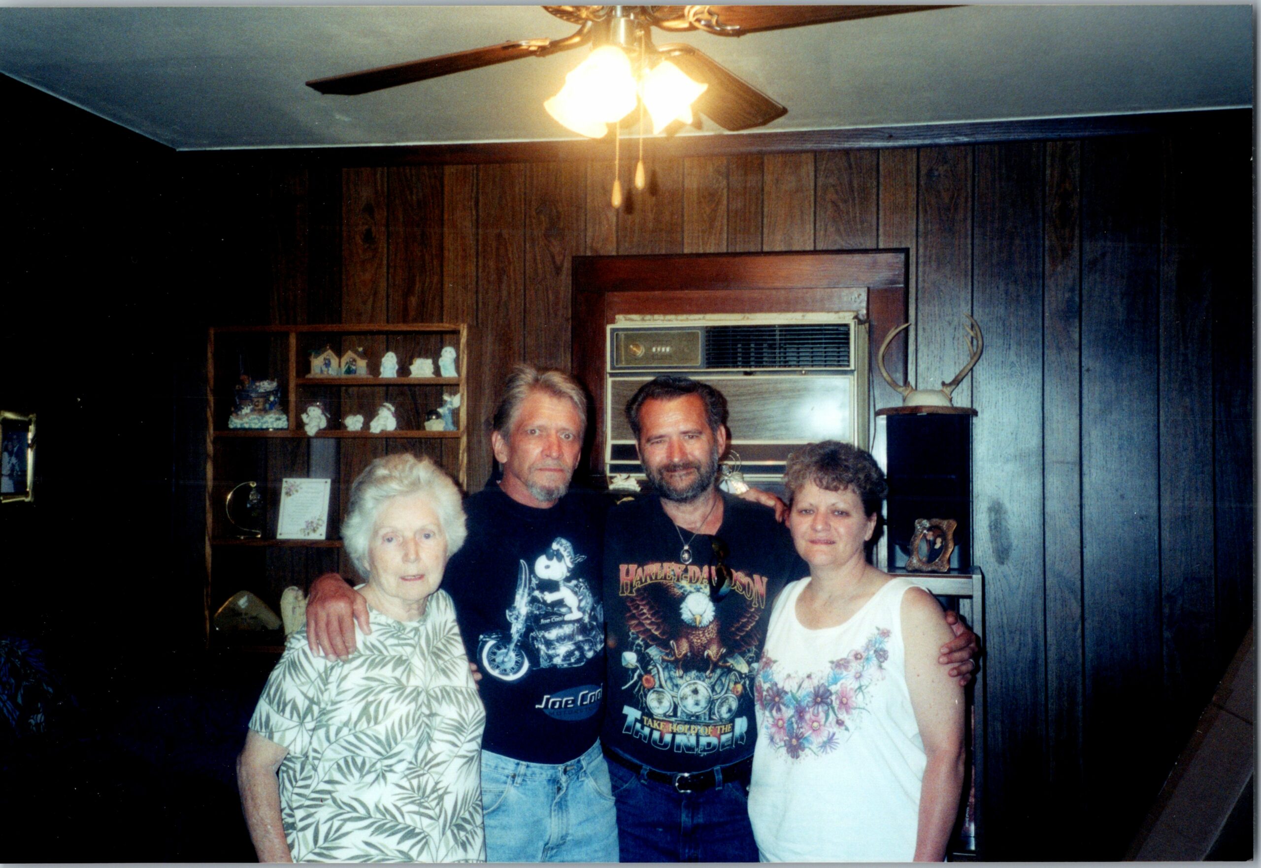 A group of five people posing together in a wood-paneled room with a ceiling fan above and a vintage cabinet in the background, looking at the camera in a casual family photo.