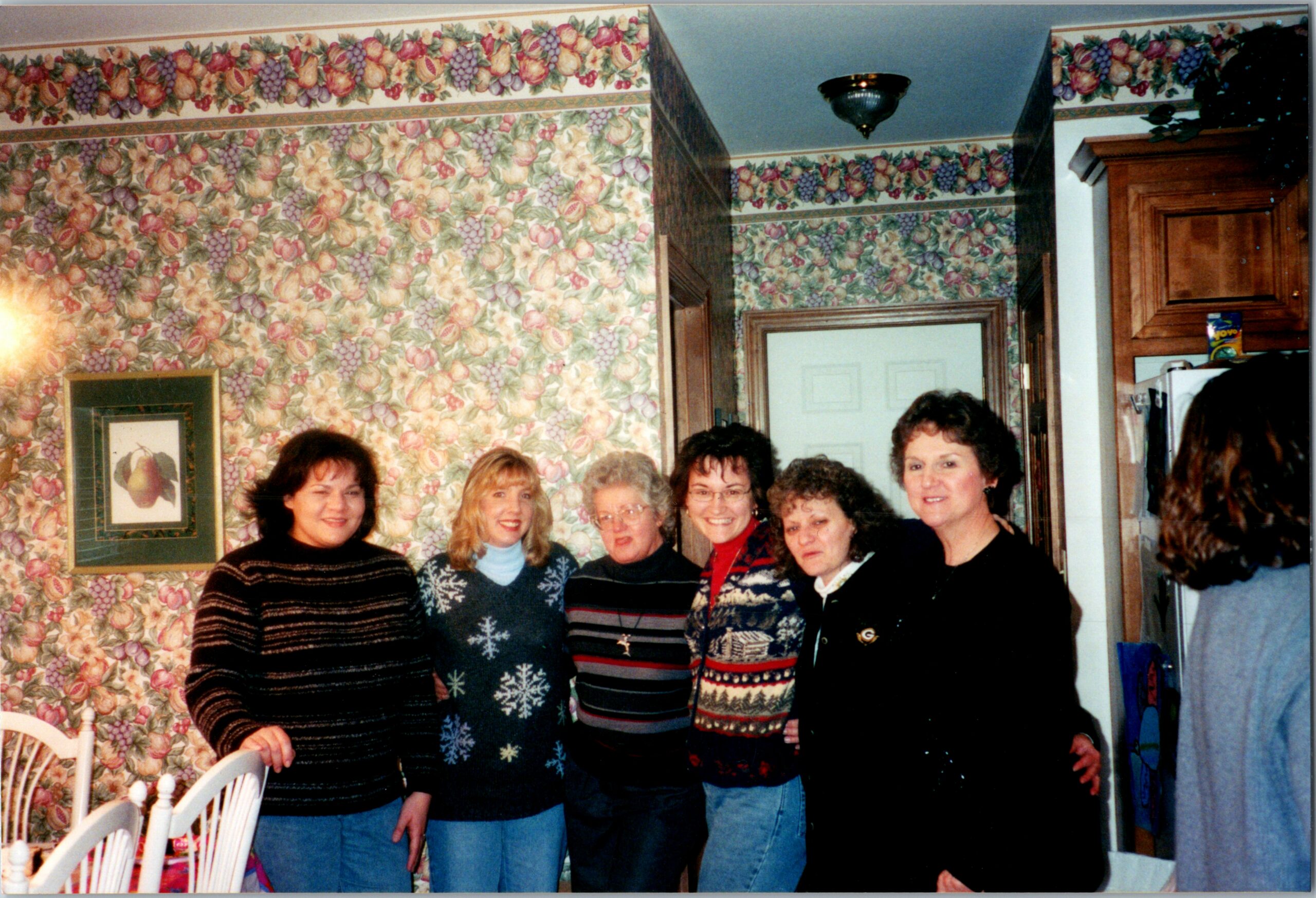 Six women in holiday sweaters posing together in a kitchen with fruit-pattern wallpaper and wooden cabinets behind them.