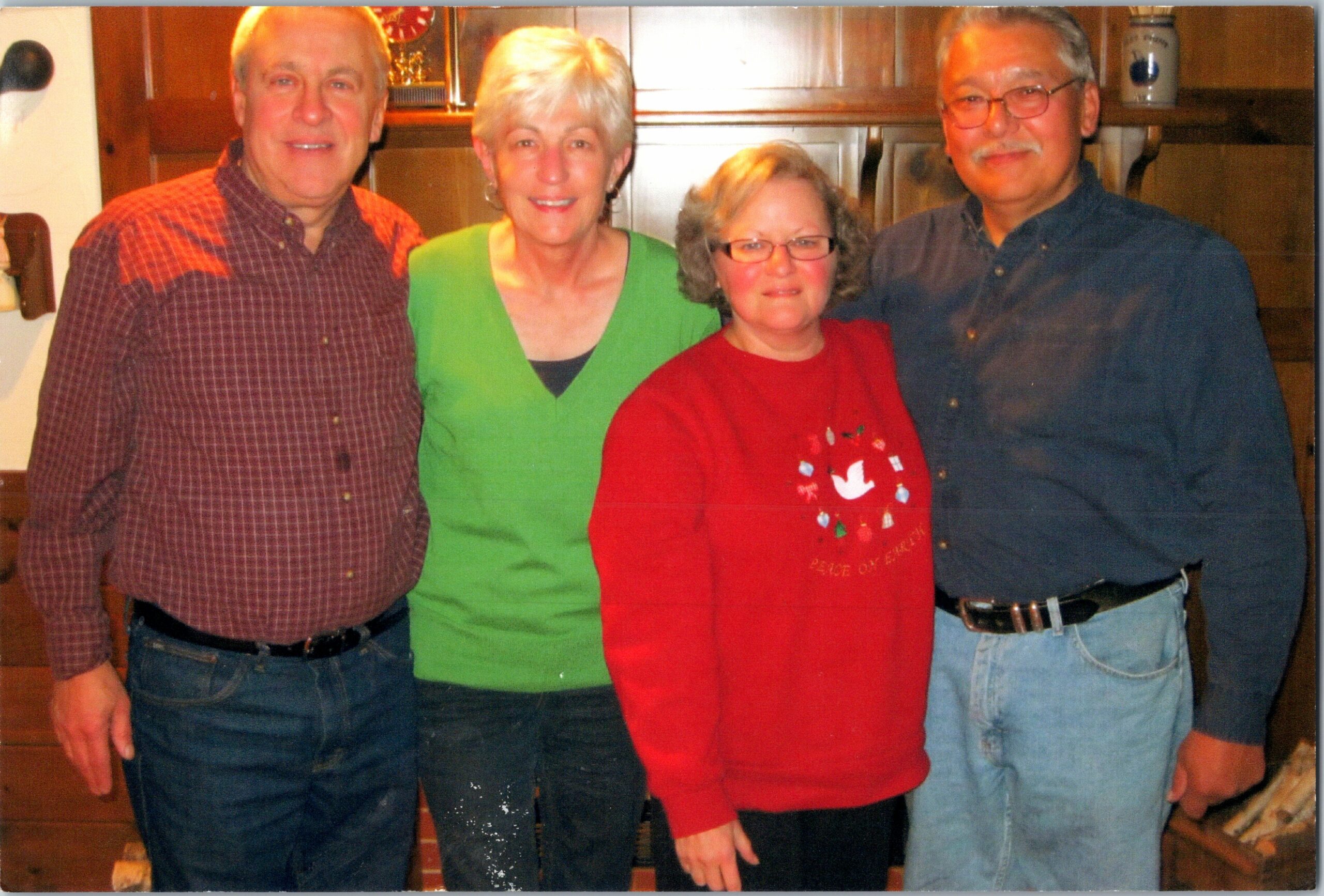 Four adults stand close together in a kitchen, smiling at the camera: man in red plaid shirt, woman in green sweater, woman in red holiday sweater, man in blue shirt.