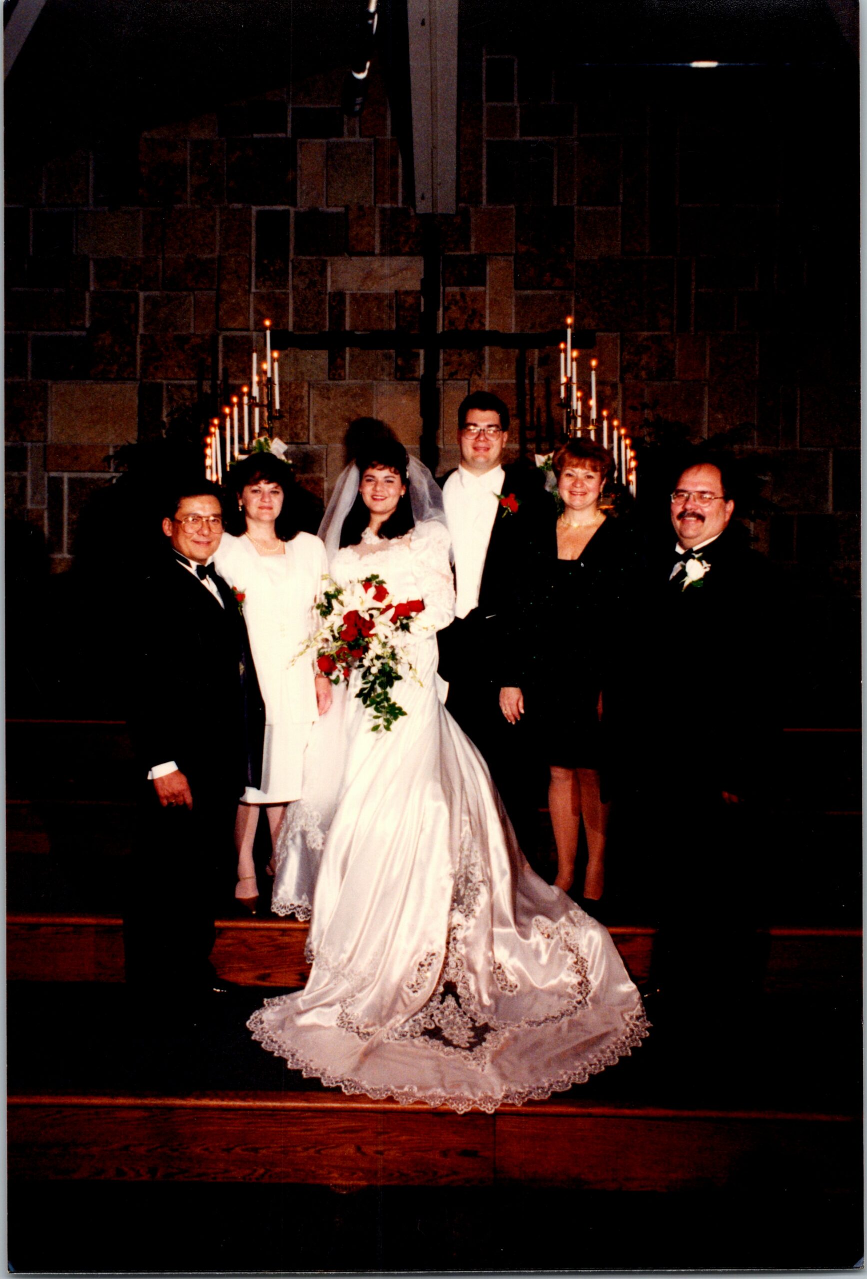 Bride and groom pose with family in a church, the bride holding a bouquet, candles glowing behind them at the altar.