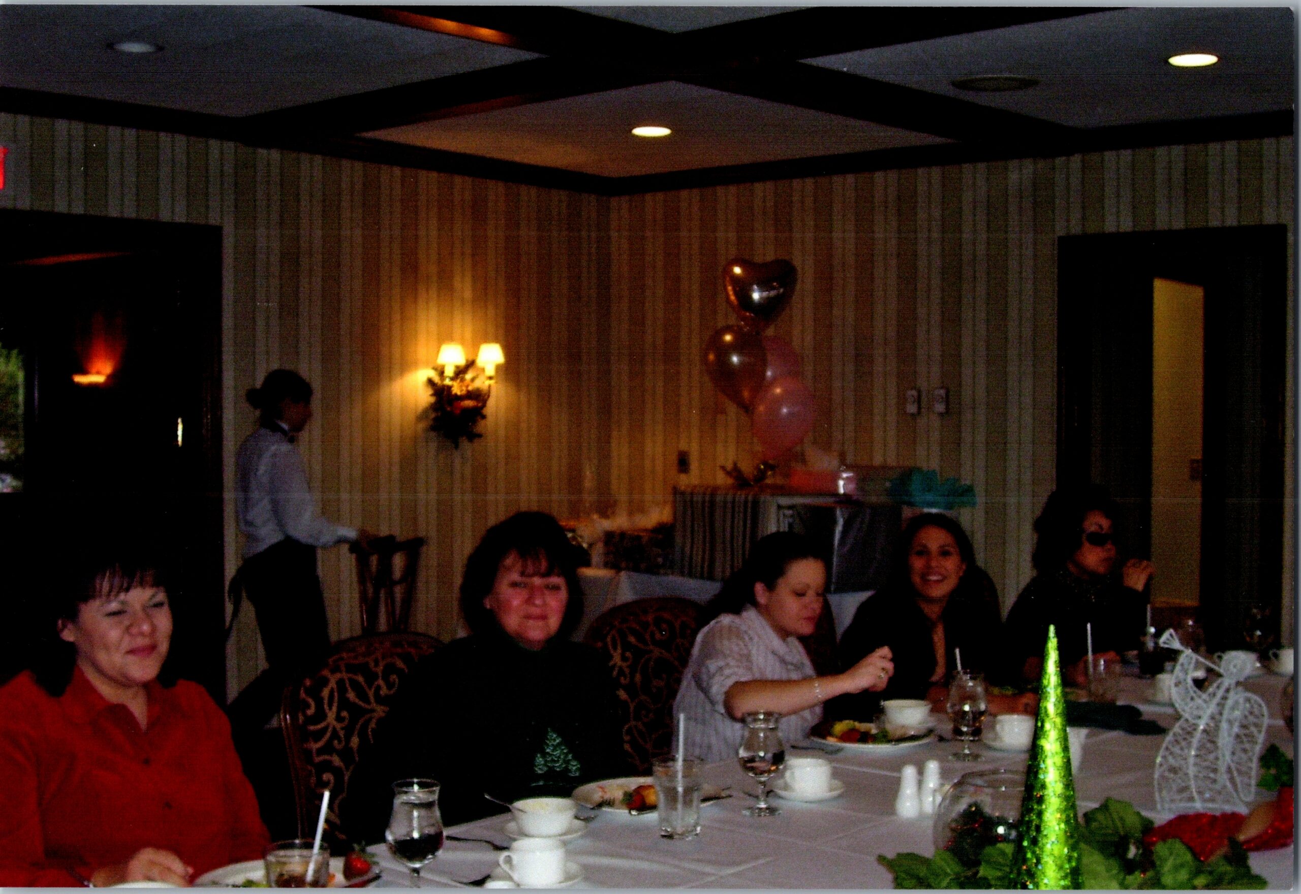 Group of people seated at a long dining table during a festive party, with balloons and celebration decor in the background.
