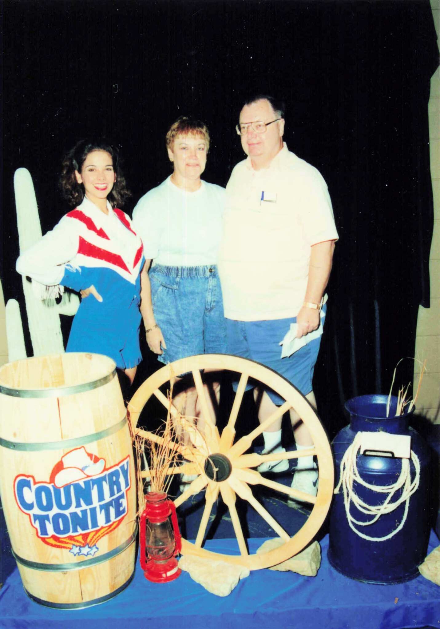 Three people posing together at a country-themed display, with a wooden barrel labeled 'Country Tonight' and a wagon wheel prop nearby.