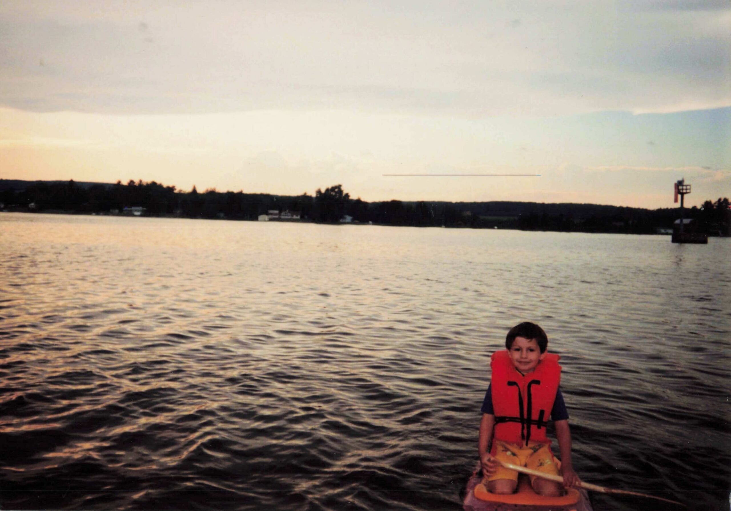 Child wearing a red life jacket sits in an orange kayak on a calm lake at sunset with a wooded shoreline in the distance.