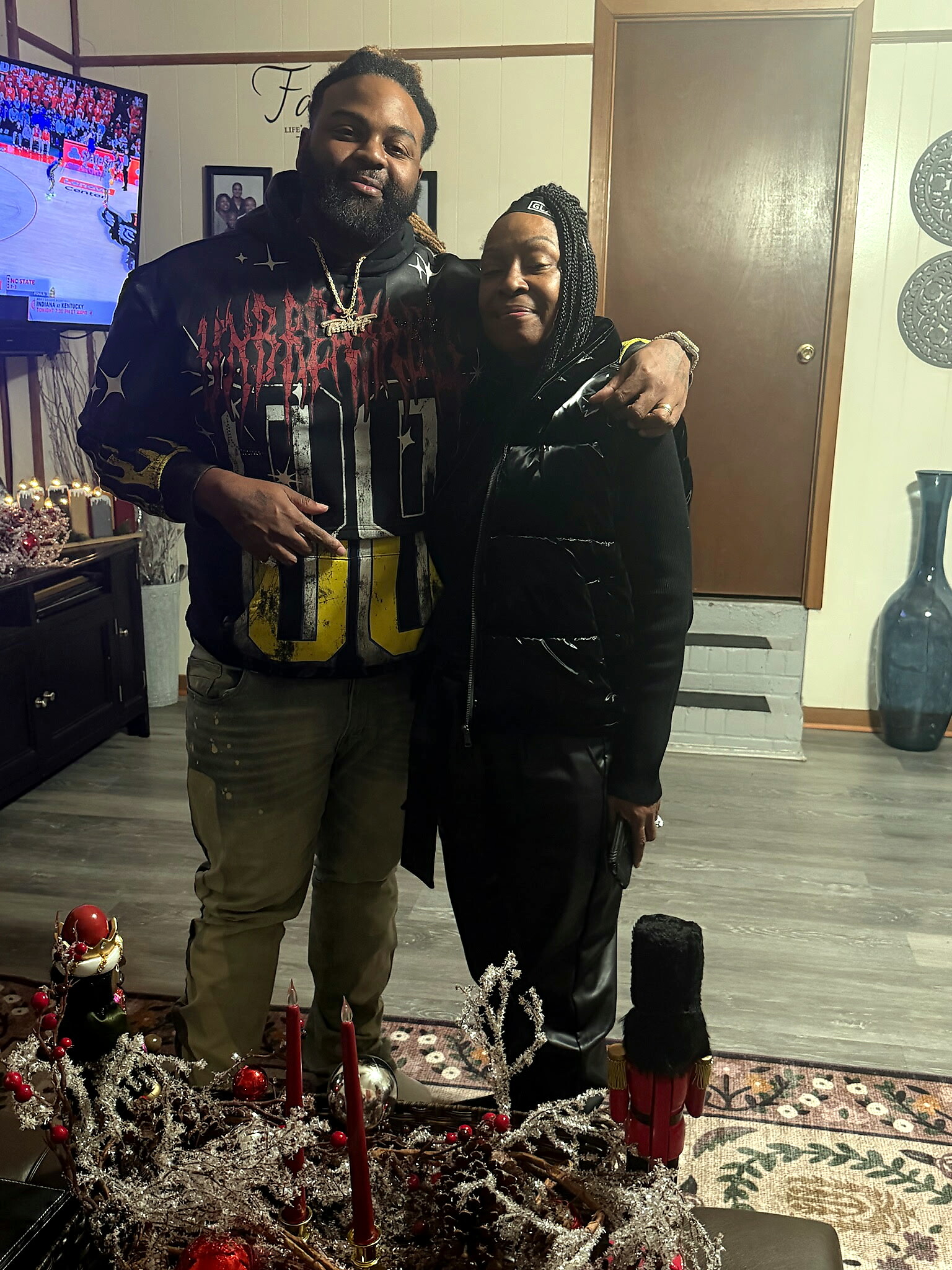 Two people pose indoors with a Christmas centerpiece in the foreground; man wearing a graphic hoodie and gold chain, woman in a black coat beside him.