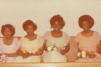 Four young girls in pastel dresses sit on a bench, holding small bouquets, posing for a family portrait.