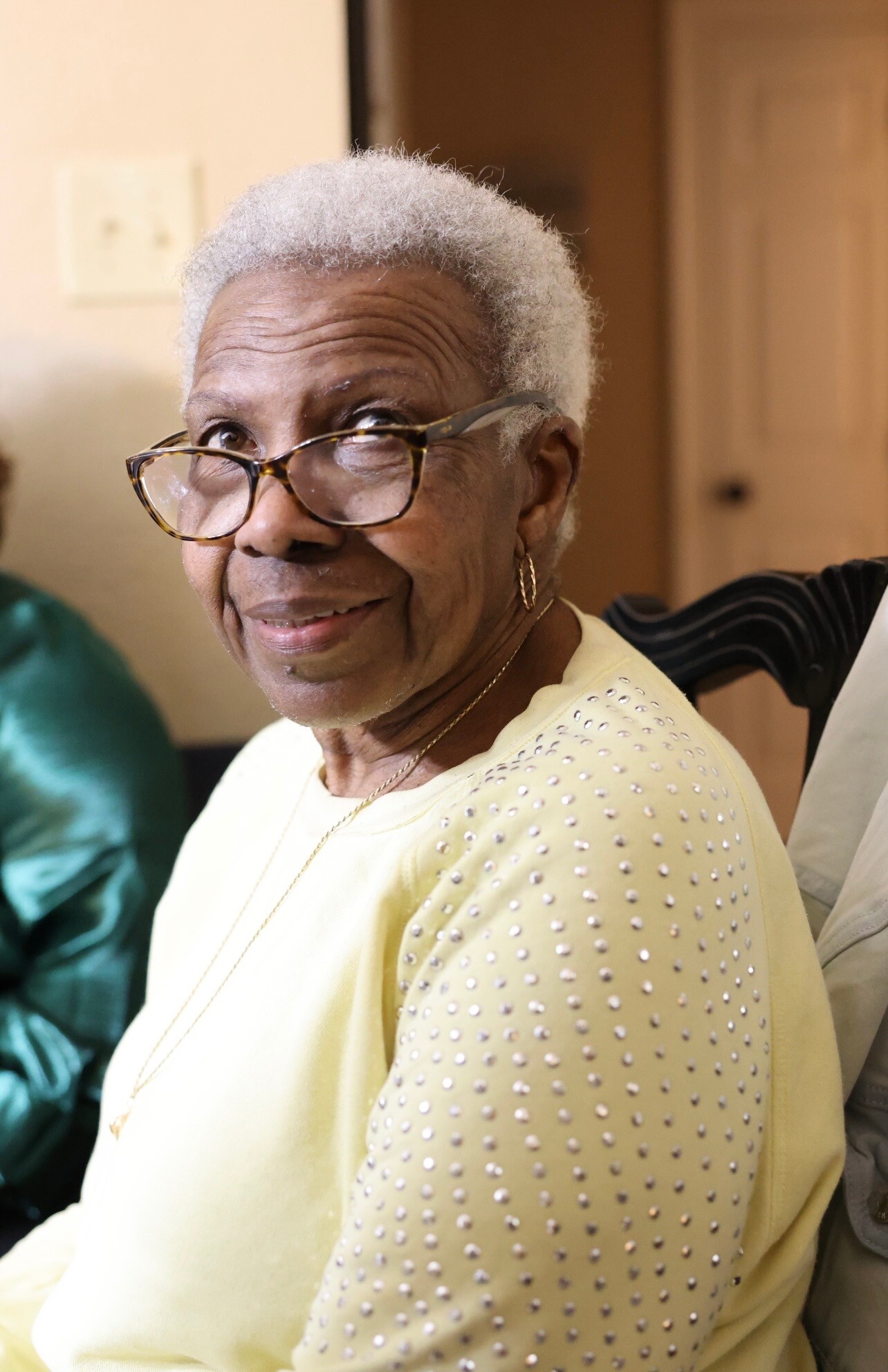 Smiling elderly woman with short gray hair and glasses wearing a pale yellow sweater with pearl-like studs, seated indoors.