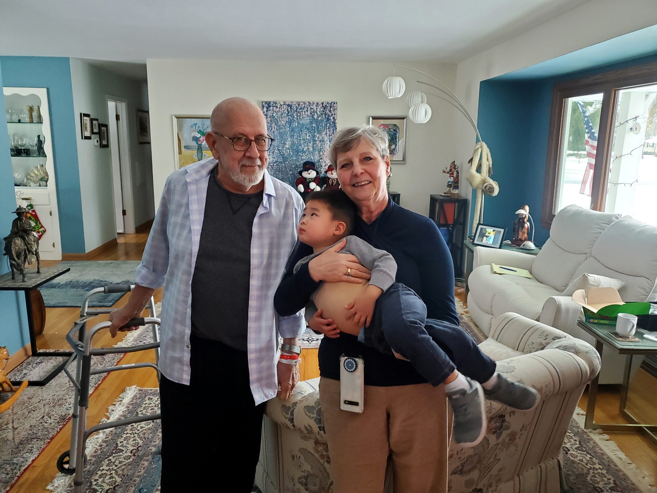 Elderly man with glasses, smiling woman, and child hugging in a cozy living room with blue walls in the background.