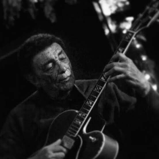 Black-and-white photo of a person playing an acoustic guitar, eyes closed, in a dim setting as they strum the instrument.