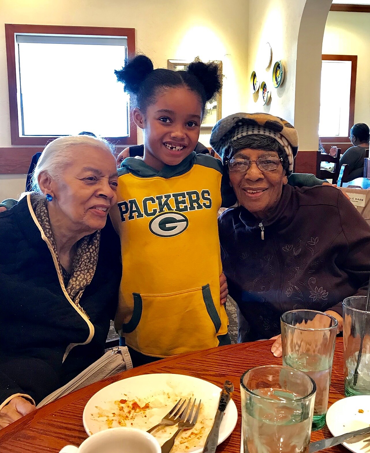A smiling young girl in a Packers hoodie stands between two elderly women at a restaurant table, with plates and glasses in front of them.