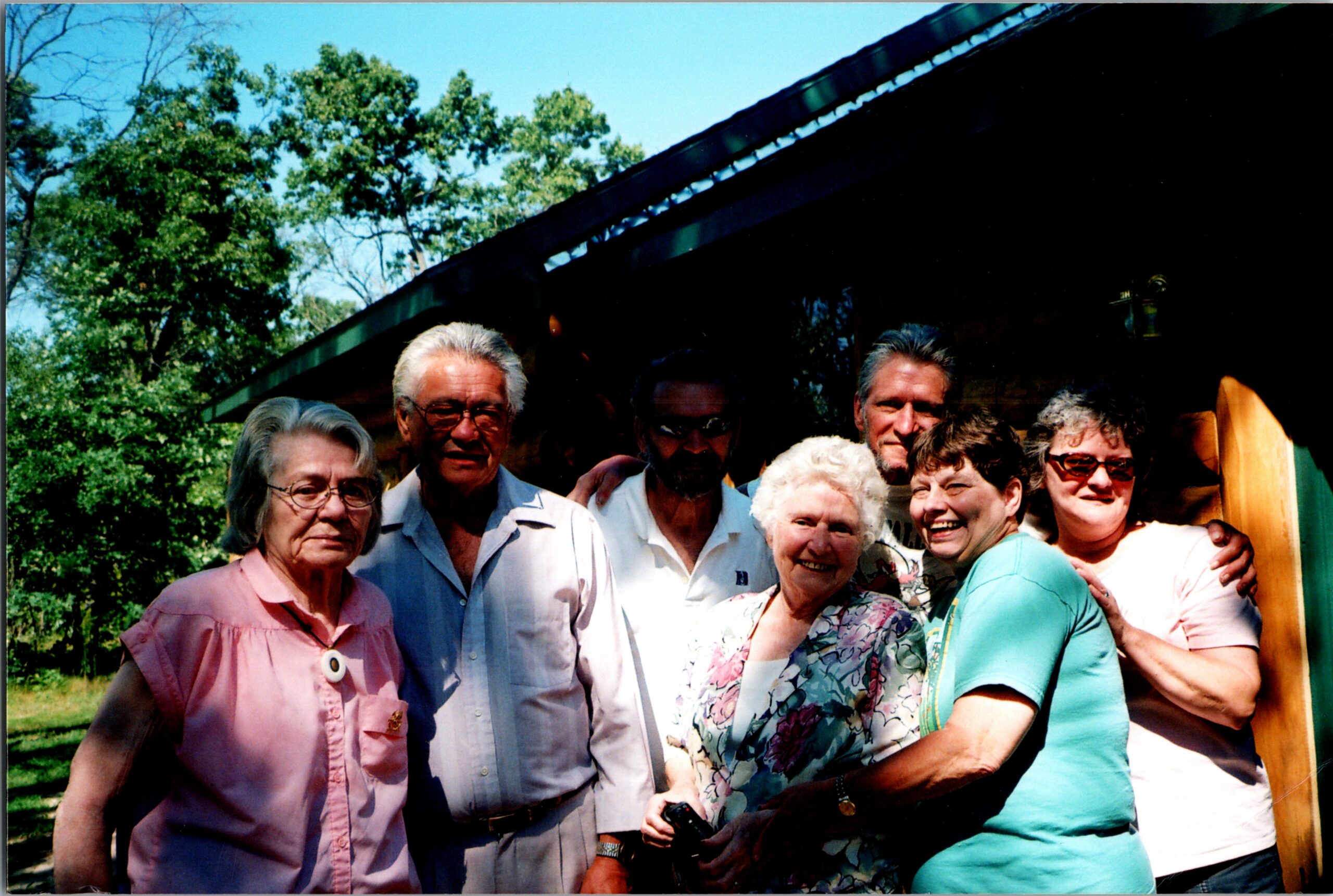 Group of smiling adults posing outdoors for a photo, standing close together in daylight with trees and a building in the background.