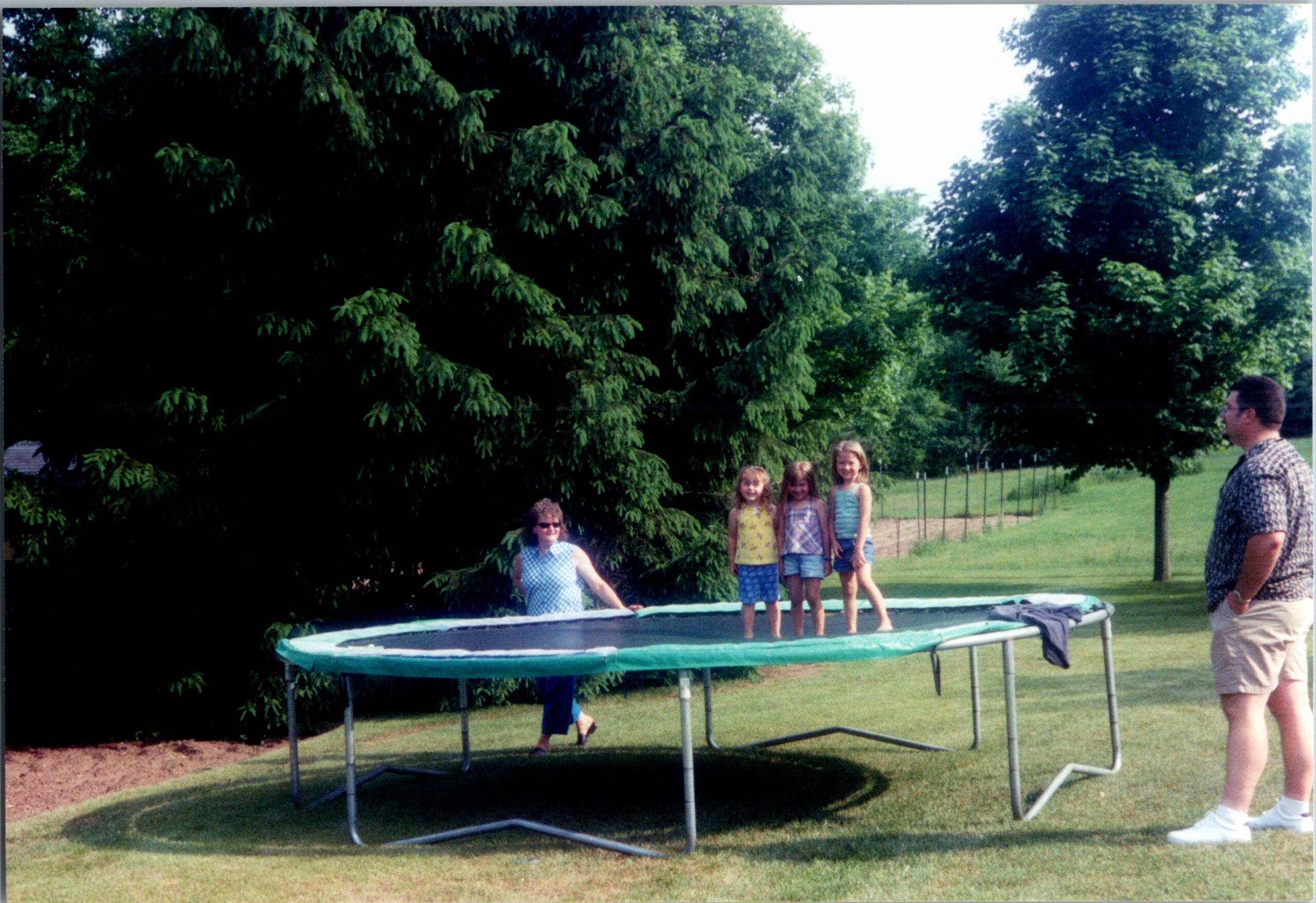 Children stand on a backyard trampoline while an adult man watches from the right; lush trees in the background.