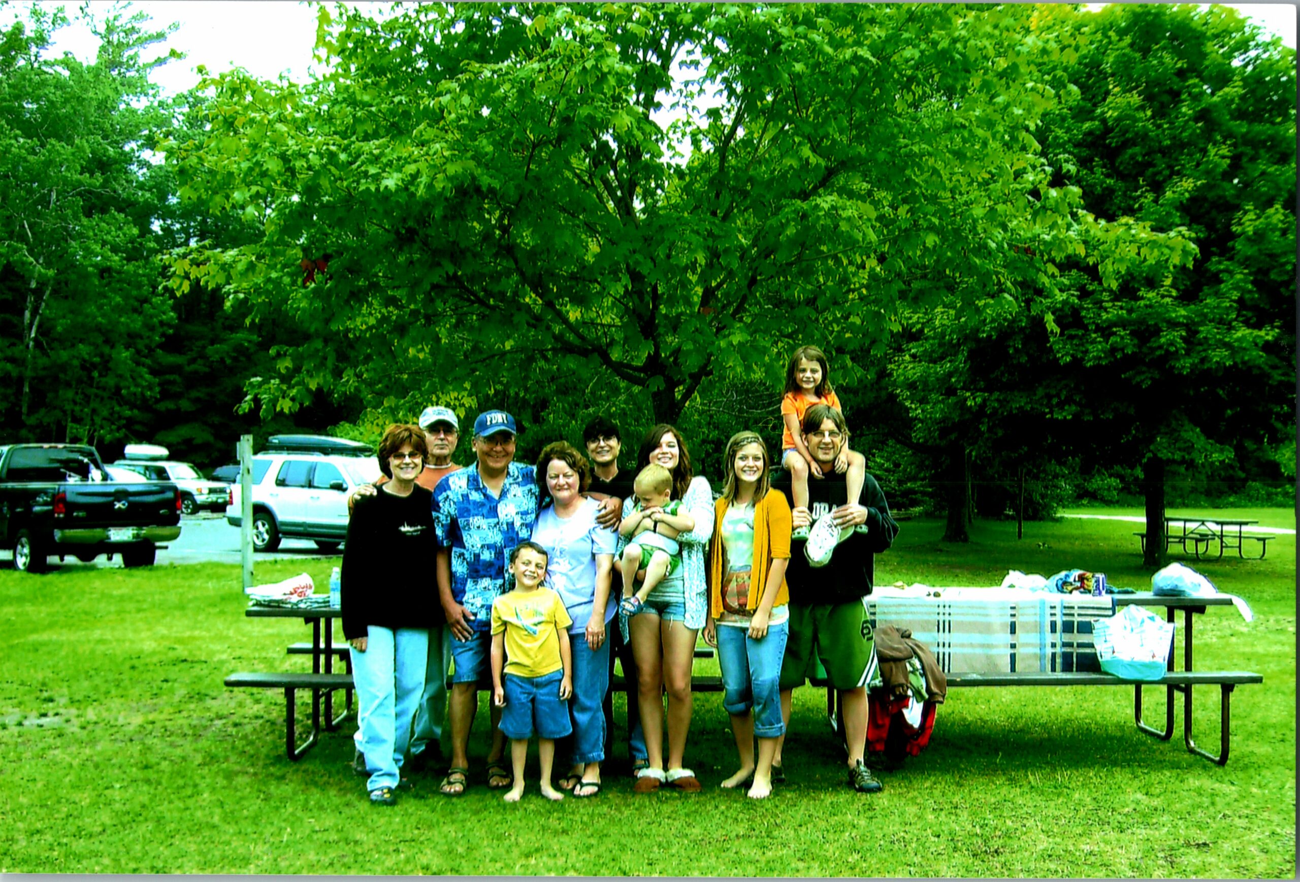 Group of family and friends posing for a photo at a park picnic area under leafy green trees on a sunny day.