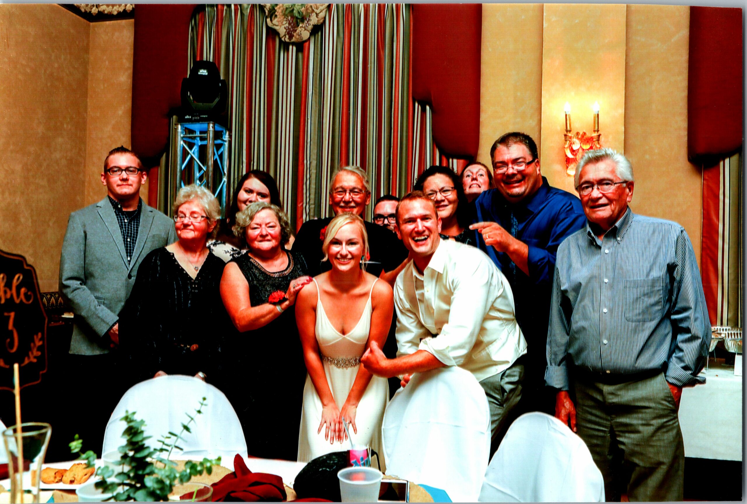 Group of smiling guests posing for a photo at a wedding reception, with the bride in a white dress front center.