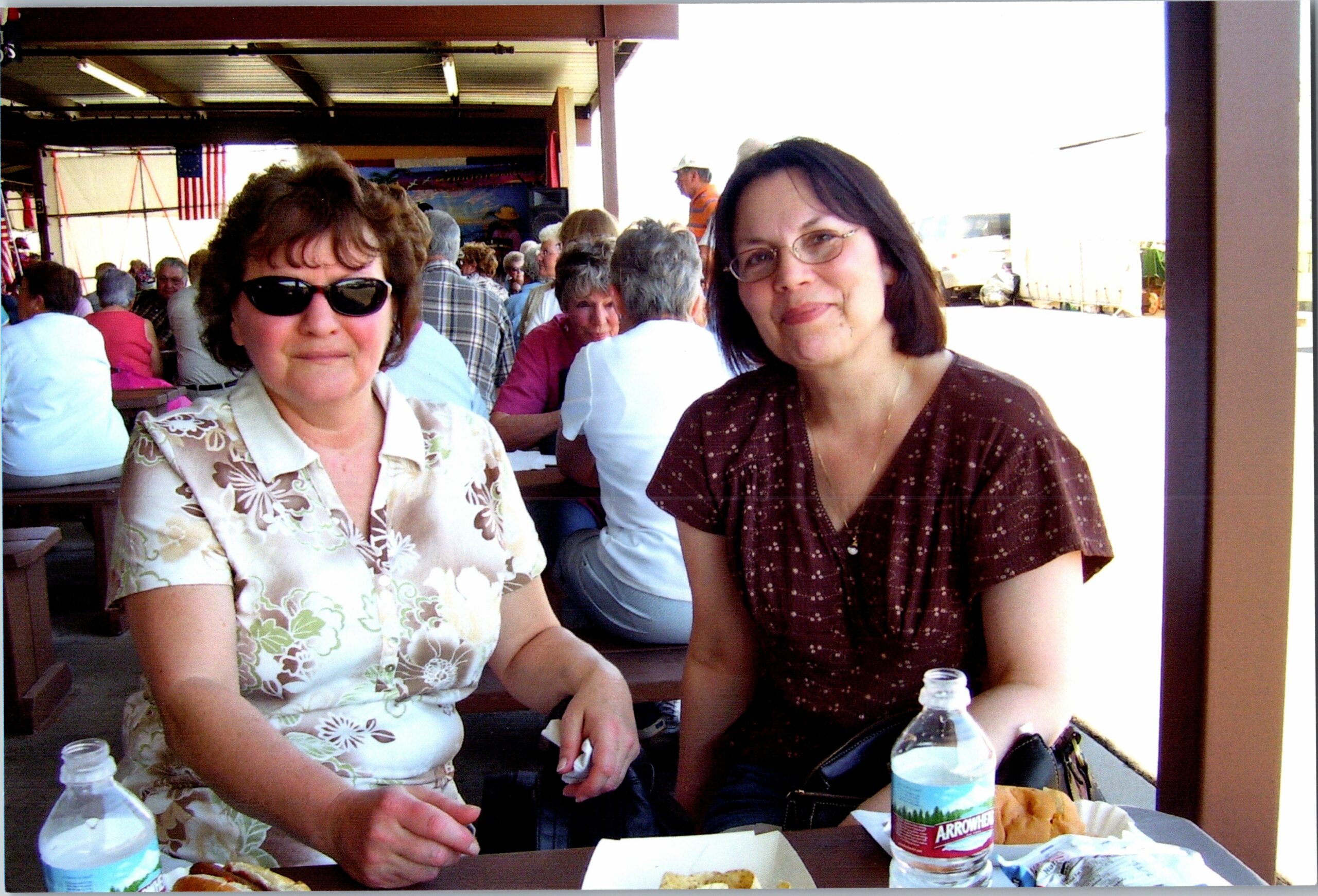 Two women sit at a picnic table during a social event, with food and drinks in front and a crowd behind them.