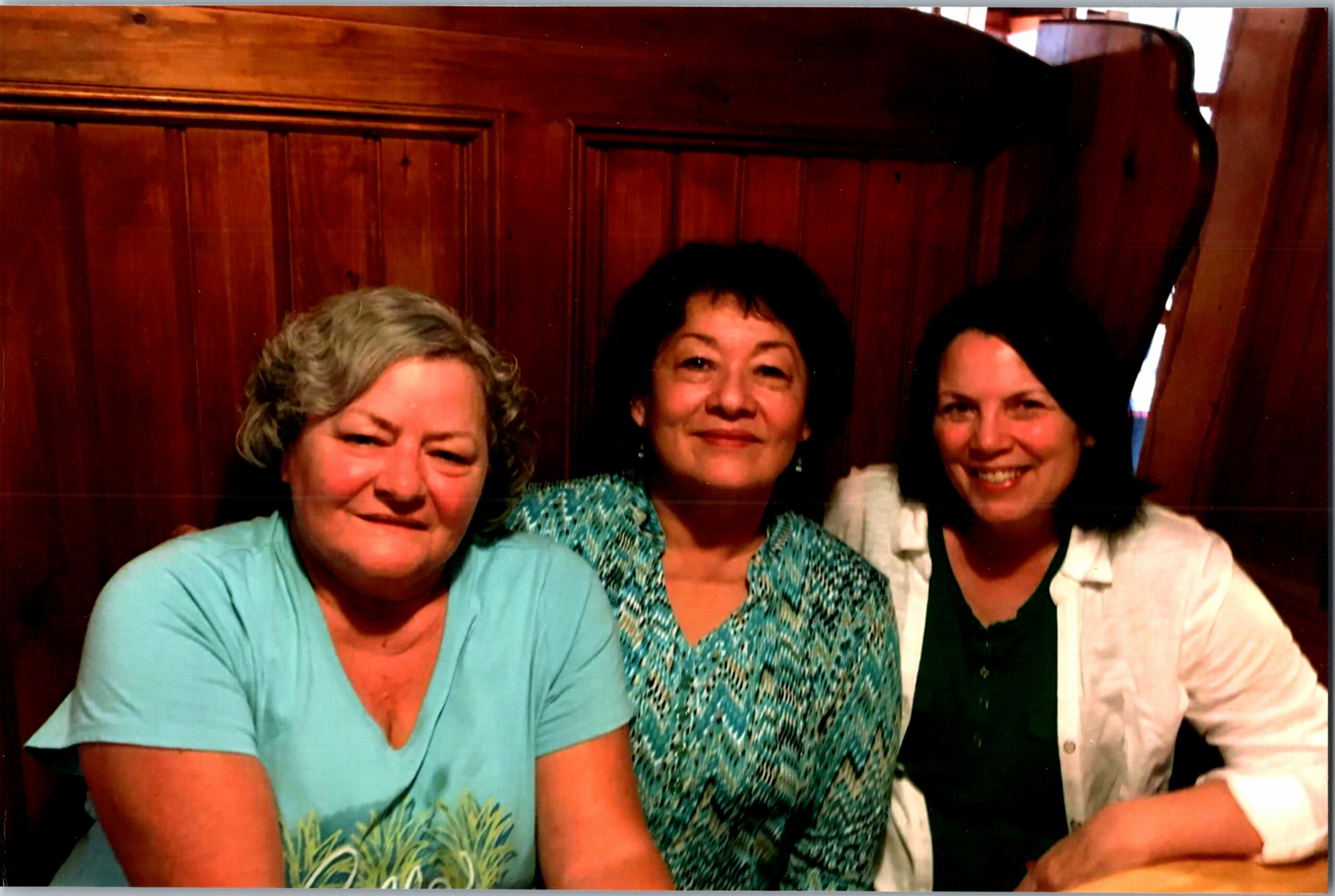 Three women smiling, seated close together indoors with a wooden paneled wall in the background.