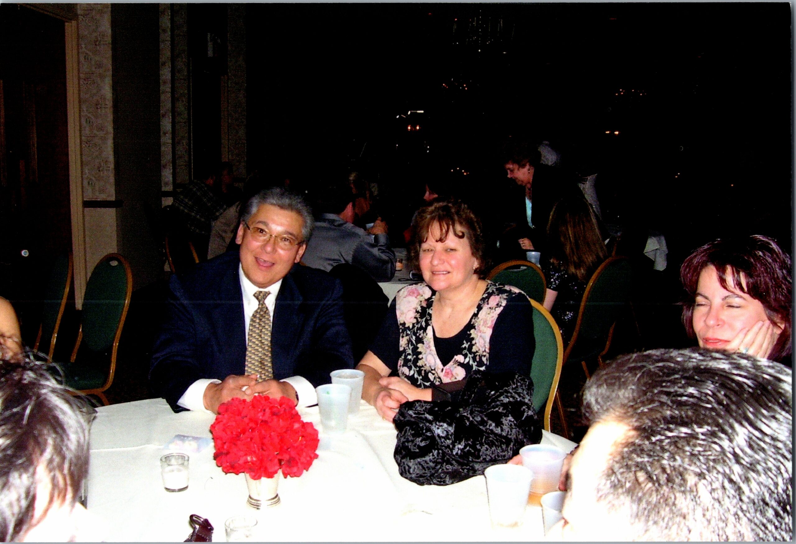 Older couple sits at a banquet table with a red flower centerpiece, smiling at a formal event.