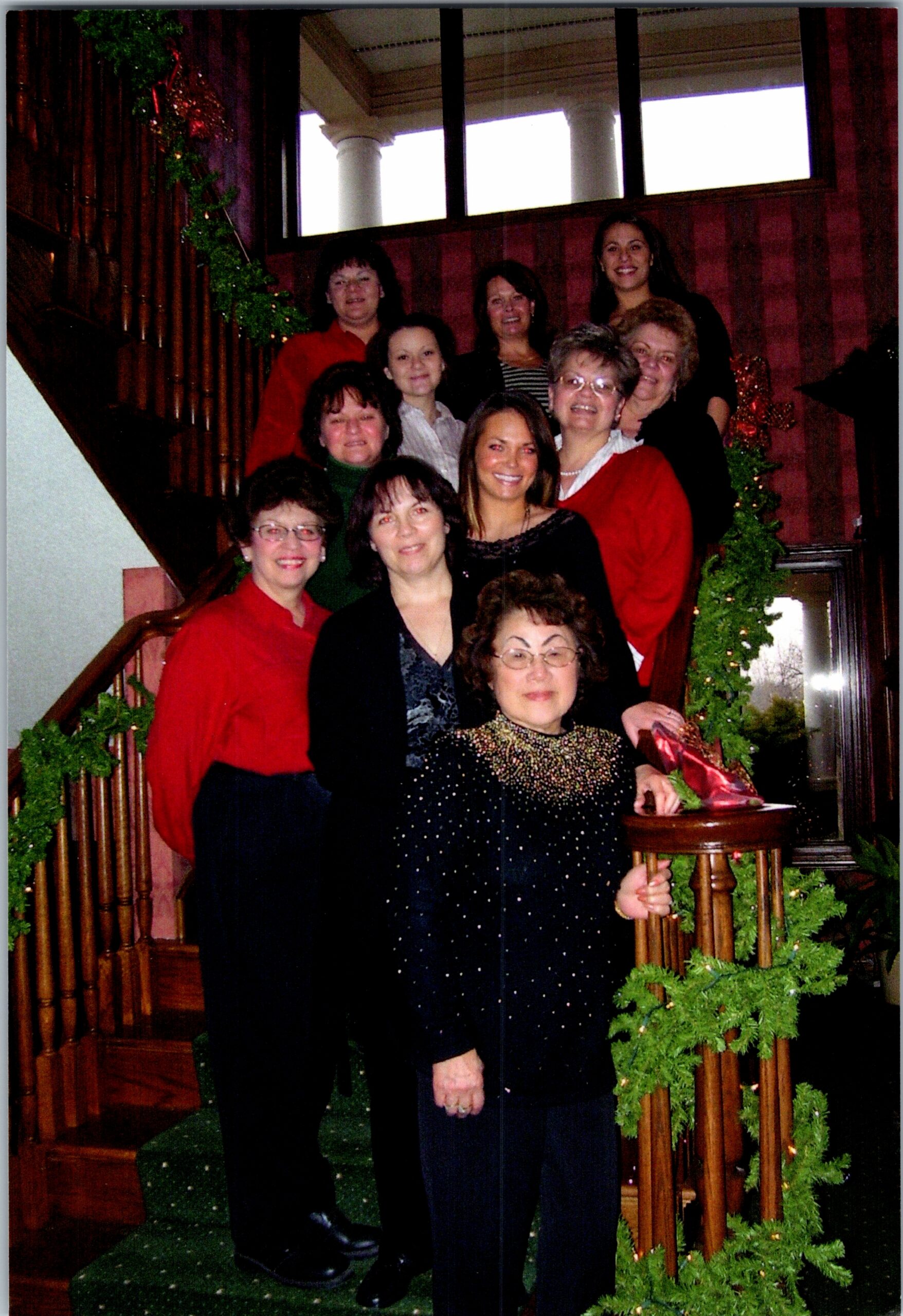 Group of women posing on a wooden staircase decorated with Christmas garlands, smiling for a photo.