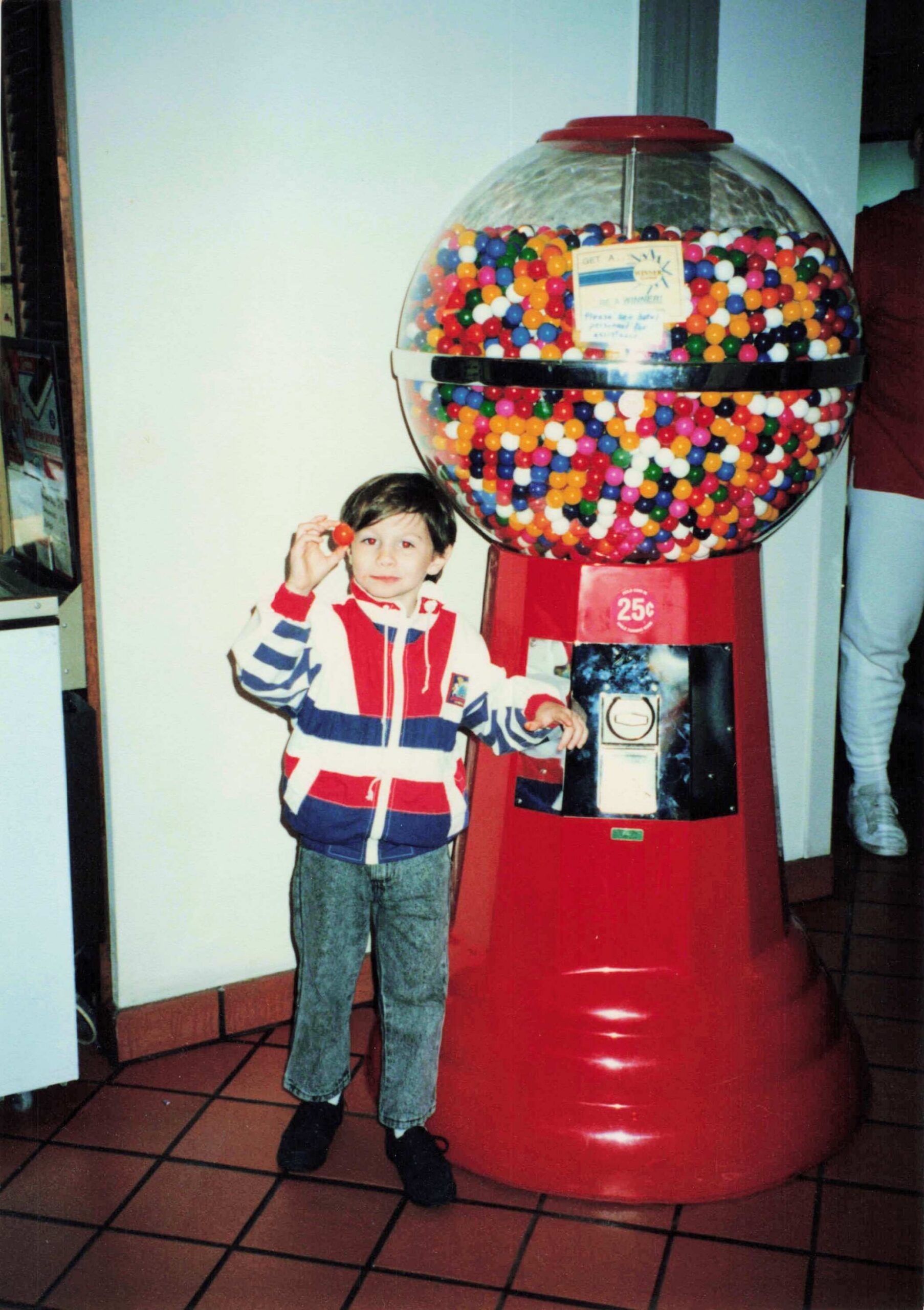 Young boy in a striped jacket holds a red gumball beside a giant transparent gumball machine filled with colorful balls.