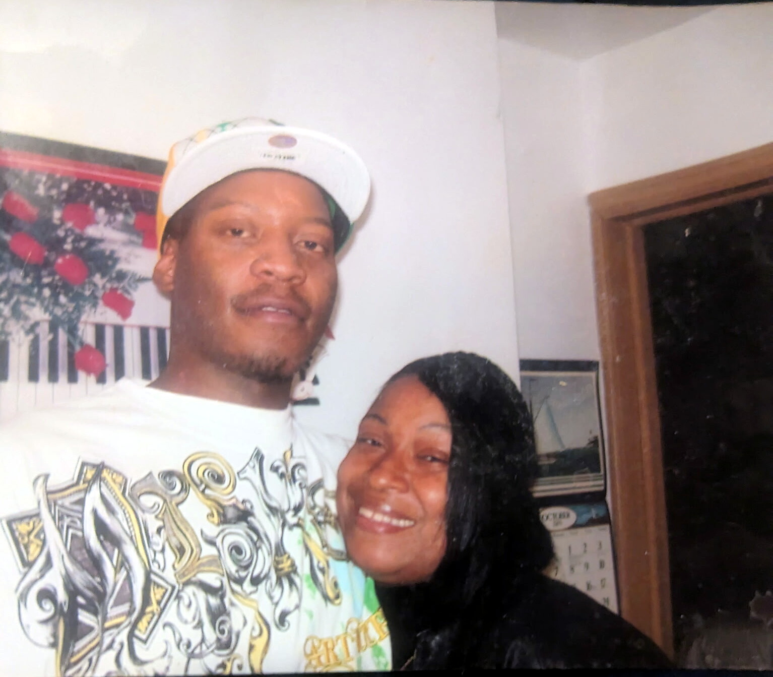 A smiling couple takes a selfie indoors; man wears a white cap and graphic shirt, woman leans in with a big smile.