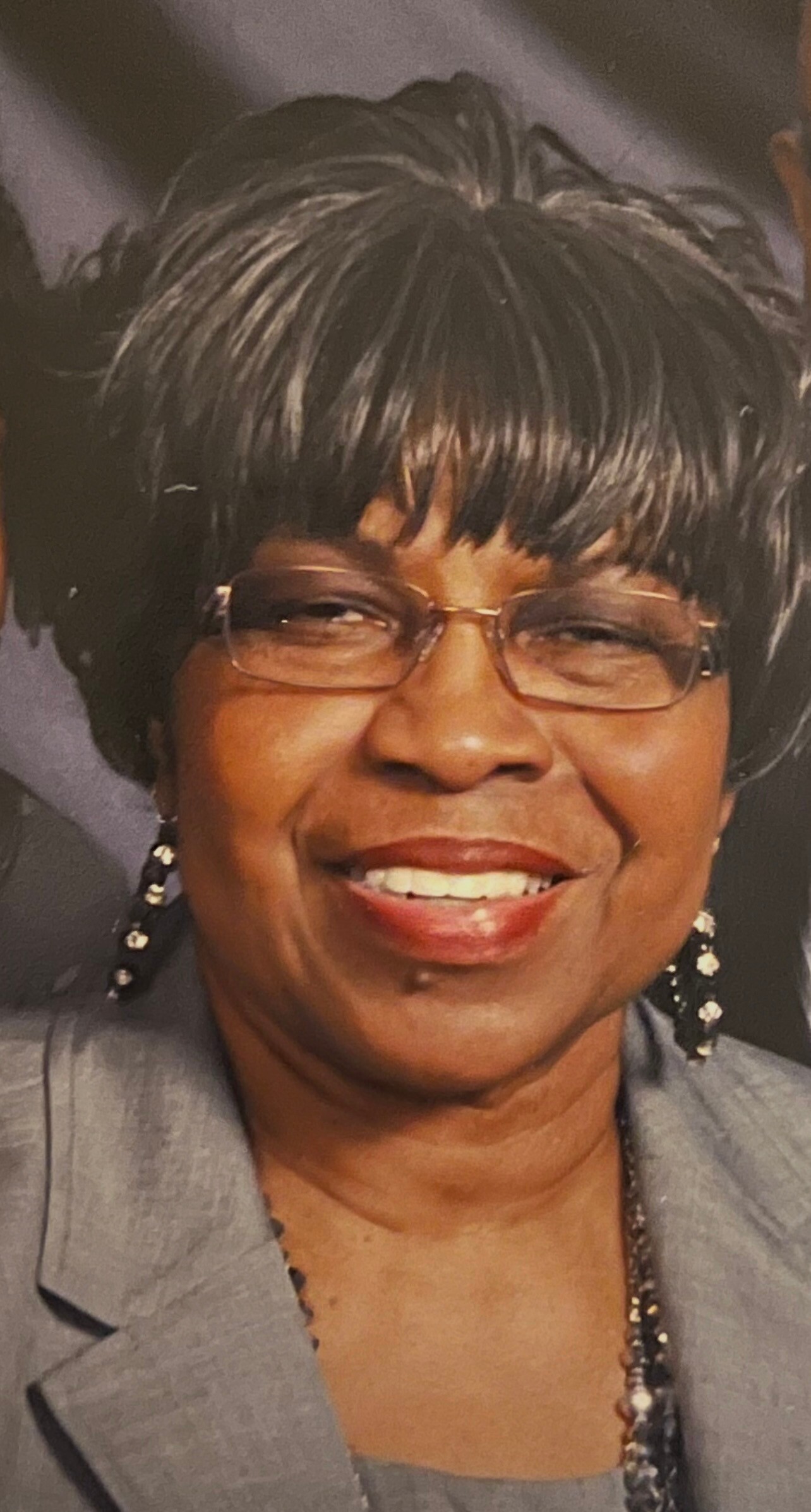 Smiling African American woman wearing glasses and a gray blazer, with short black and gray hair and dangling earrings.
